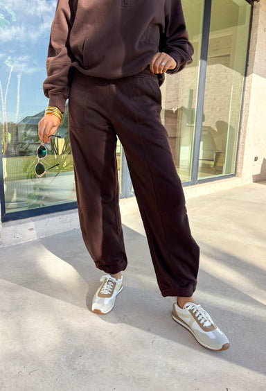 Stay cozy in style with these chocolate brown barrel joggers by Rae Mode. Lined in soft fleece and finished with a drawstring elastic waistband, these joggers offer the perfect blend of comfort and warmth for chilly winter days. Featuring side and back pockets for everyday convenience, their relaxed barrel silhouette makes them ideal for lounging or layering on the go. Pair them with the matching quarter-zip pullover for a complete cozy set or wear them solo for a laid-back, stylish look.