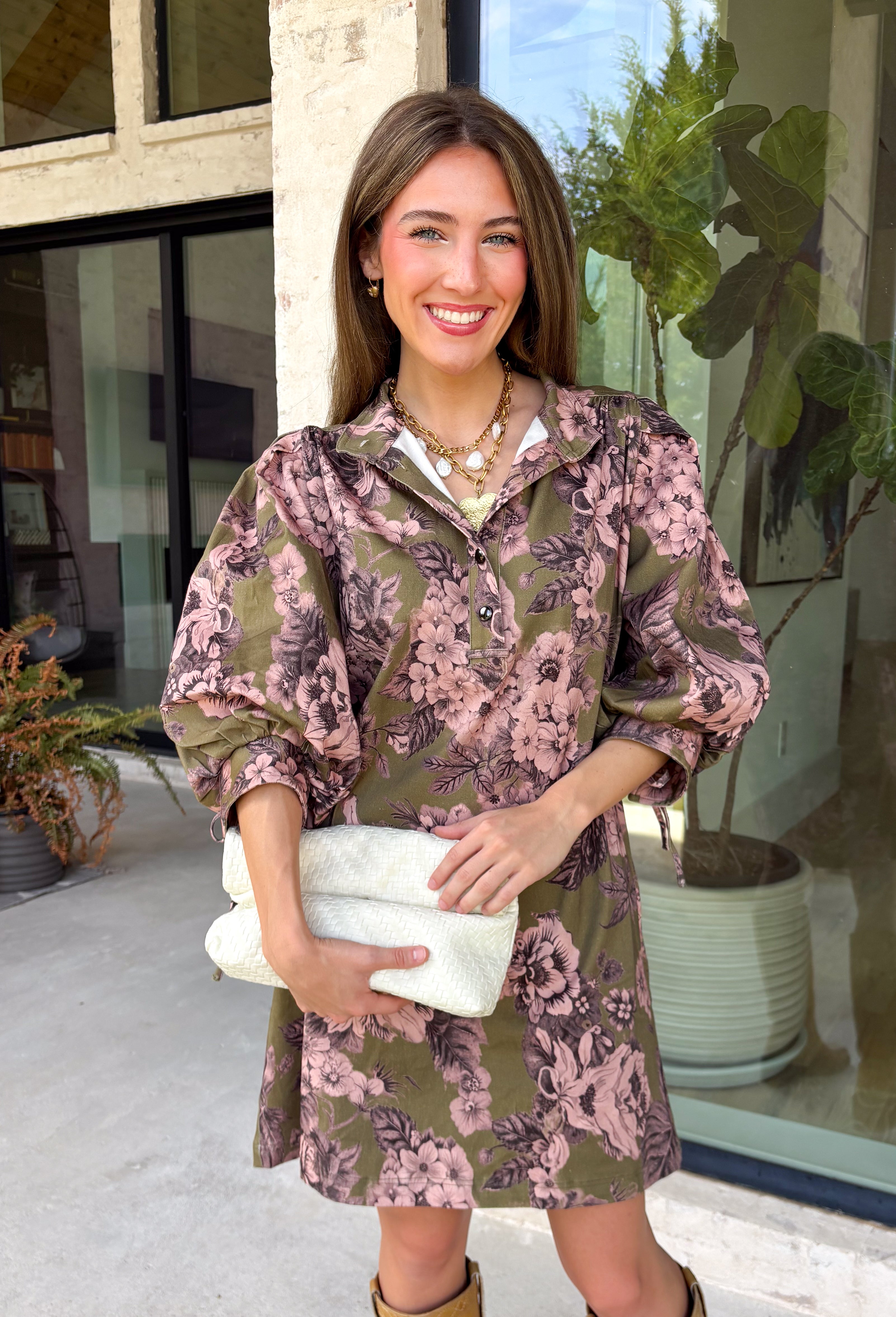 Bring effortless charm to your fall wardrobe with this beautifully detailed dress from Entro. Designed in a rich olive hue with a delicate floral print in shades of light pink and black, this timeless dress is perfect for everything from brunch dates to weekend outings. The stand neckline and partial button-front closure create a polished silhouette, while the ¾ puff sleeves are finished with sweet tie cuffs for an extra feminine touch. 