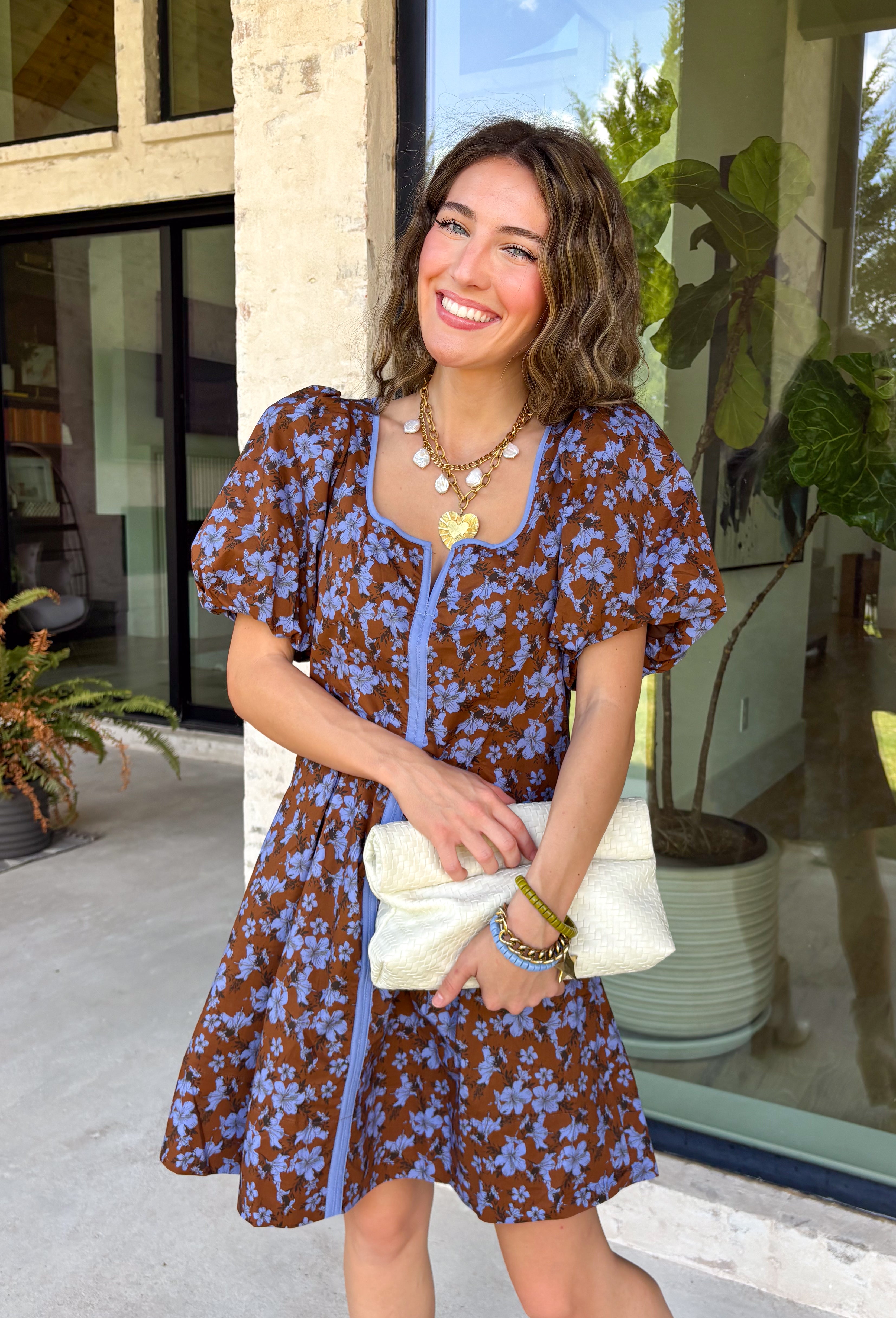 Bring timeless elegance to your fall wardrobe with this beautifully detailed dress from Entro. Designed with a rich brown backdrop and a stunning blue floral print, this feminine dress is full of eye-catching seasonal charm. The flattering square neckline is accented with beautiful blue contrast trim, while the pleated waist creates a flattering, defined silhouette. 