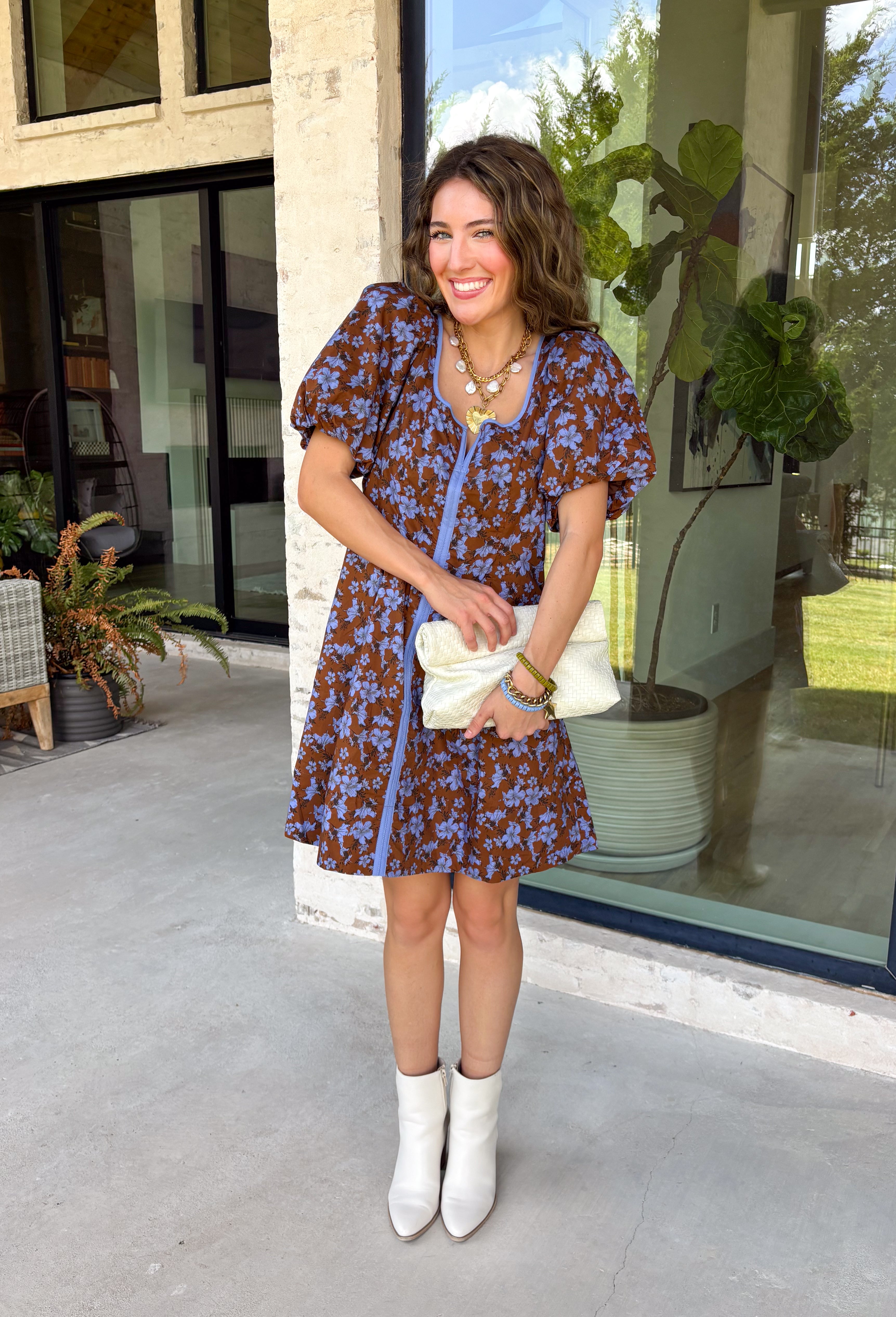 Bring timeless elegance to your fall wardrobe with this beautifully detailed dress from Entro. Designed with a rich brown backdrop and a stunning blue floral print, this feminine dress is full of eye-catching seasonal charm. The flattering square neckline is accented with beautiful blue contrast trim, while the pleated waist creates a flattering, defined silhouette. 