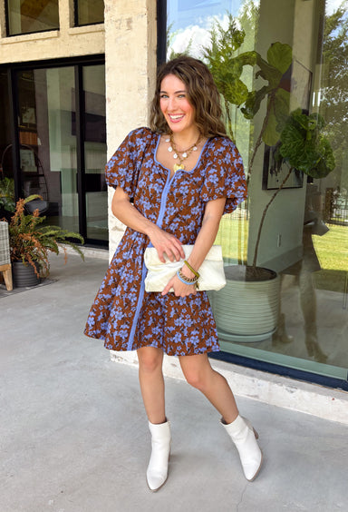 Bring timeless elegance to your fall wardrobe with this beautifully detailed dress from Entro. Designed with a rich brown backdrop and a stunning blue floral print, this feminine dress is full of eye-catching seasonal charm. The flattering square neckline is accented with beautiful blue contrast trim, while the pleated waist creates a flattering, defined silhouette. 