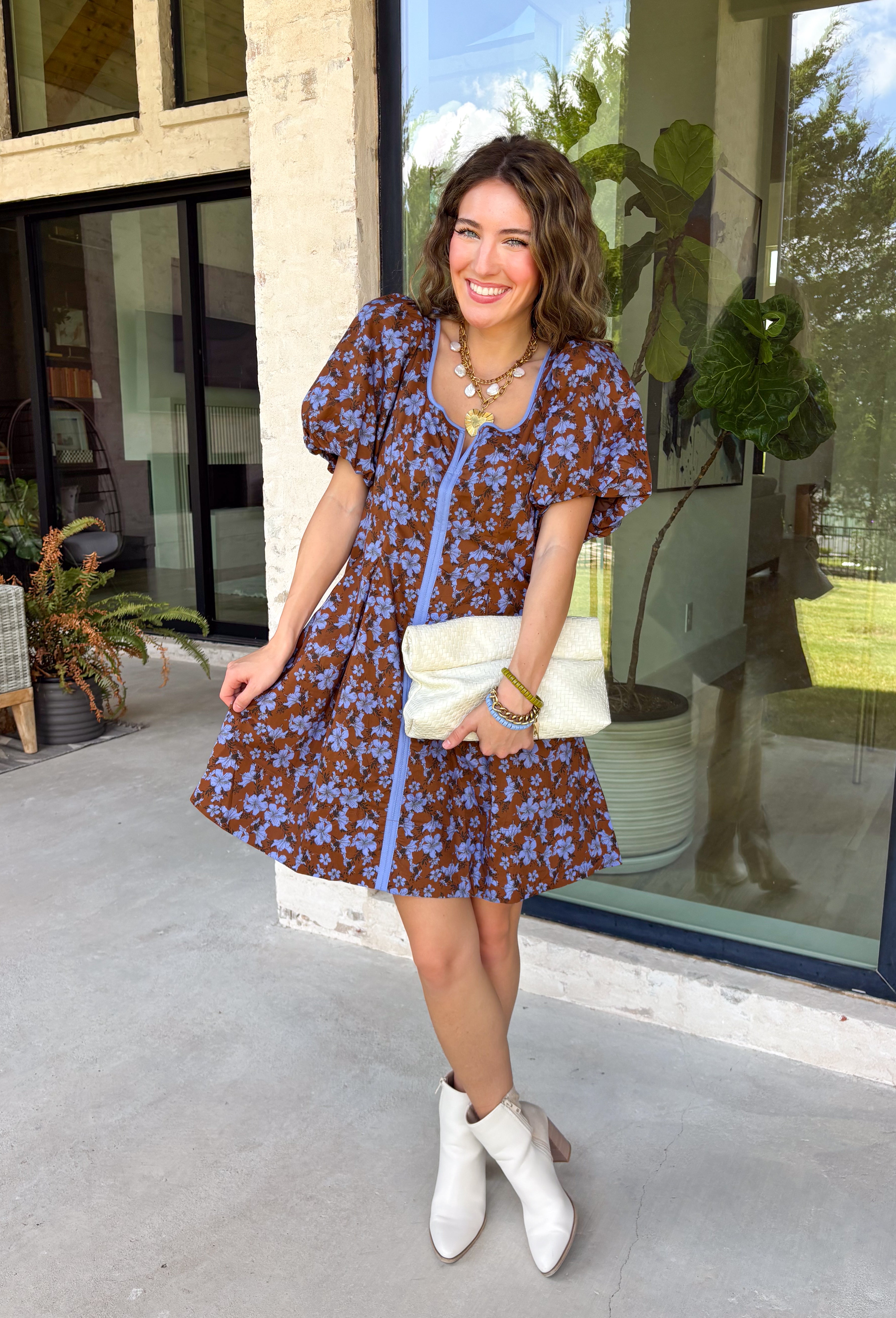 Bring timeless elegance to your fall wardrobe with this beautifully detailed dress from Entro. Designed with a rich brown backdrop and a stunning blue floral print, this feminine dress is full of eye-catching seasonal charm. The flattering square neckline is accented with beautiful blue contrast trim, while the pleated waist creates a flattering, defined silhouette. 