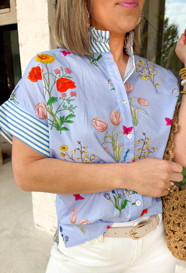 Bright, cheerful, and full of spring personality, this light blue button-down top by Entro is an instant outfit-lifter. Featuring a colorful floral print with pops of pink, green, red, and yellow, it brings a playful feel to a classic silhouette. The blue and white striped sleeves and neckline add a fun contrast, while the subtle ruffle neckline gives it a soft, feminine finish. Lightweight cotton makes it perfect for warm days and easy layering.
