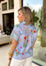 Bright, cheerful, and full of spring personality, this light blue button-down top by Entro is an instant outfit-lifter. Featuring a colorful floral print with pops of pink, green, red, and yellow, it brings a playful feel to a classic silhouette. The blue and white striped sleeves and neckline add a fun contrast, while the subtle ruffle neckline gives it a soft, feminine finish. Lightweight cotton makes it perfect for warm days and easy layering.
