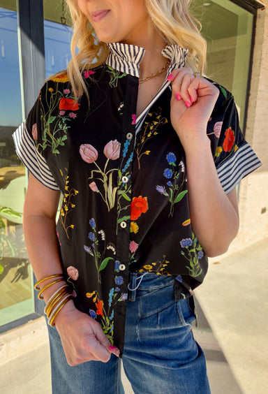 Bright, cheerful, and full of spring personality, this black button-down top by Entro is an instant outfit-lifter. Featuring a colorful floral print with pops of pink, green, red, blue, and yellow, it brings a playful feel to a classic silhouette. The striped sleeves and neckline add a fun contrast, while the subtle ruffle neckline gives it a soft, feminine finish. Lightweight cotton makes it perfect for warm days and easy layering.