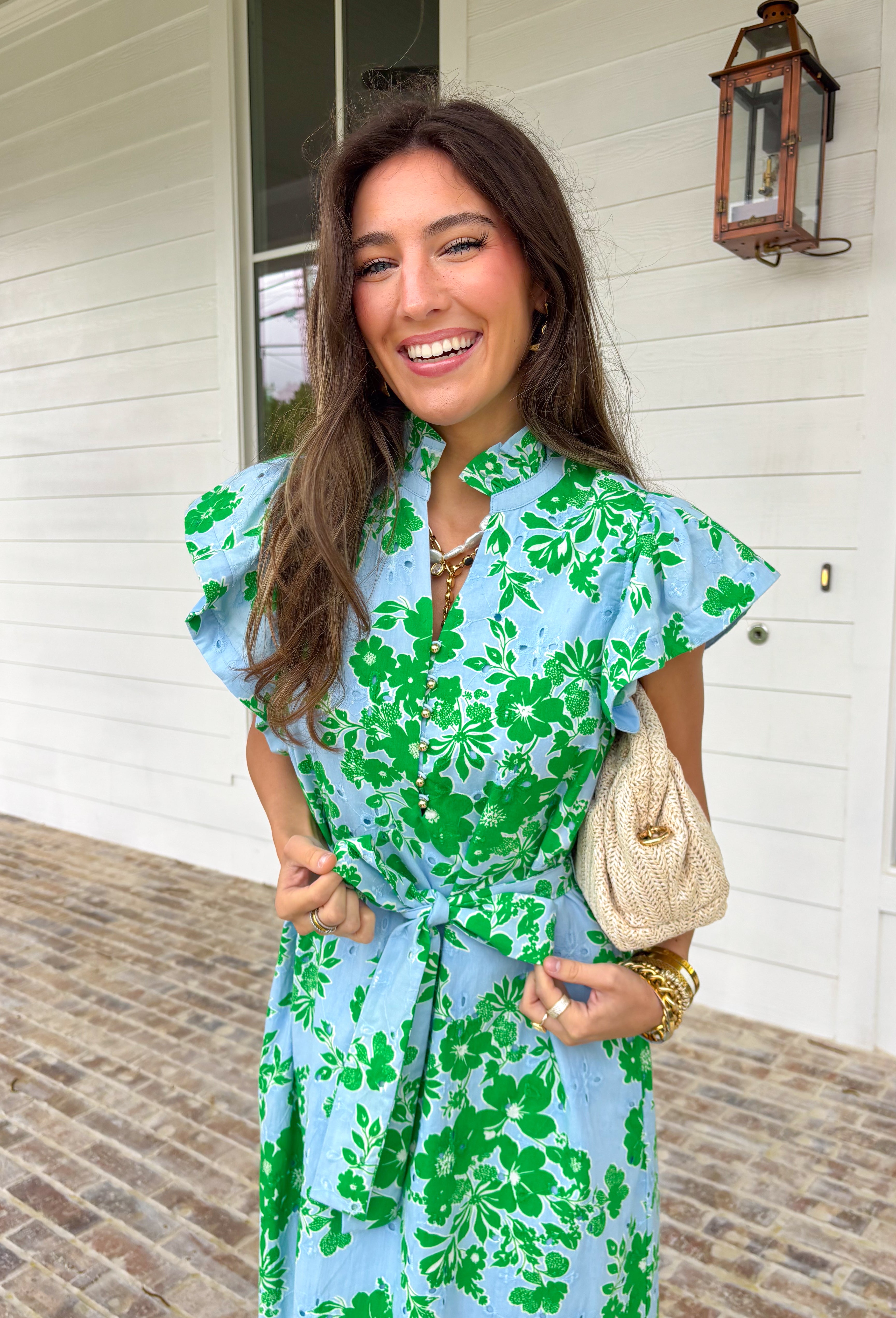 Soft, feminine, and full of spring charm! This light blue midi dress features delicate eyelet lace detailing throughout paired with a fresh green floral print for the prettiest layered texture. The subtle ruffle neckline adds a romantic touch, while the gold button details down the front bring a hint of shine. Designed with a matching self-tie closure to gently define the waist and a flowy tiered skirt for beautiful movement, this dress feels both polished and easy to wear. 