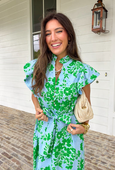 Soft, feminine, and full of spring charm! This light blue midi dress features delicate eyelet lace detailing throughout paired with a fresh green floral print for the prettiest layered texture. The subtle ruffle neckline adds a romantic touch, while the gold button details down the front bring a hint of shine. Designed with a matching self-tie closure to gently define the waist and a flowy tiered skirt for beautiful movement, this dress feels both polished and easy to wear. 
