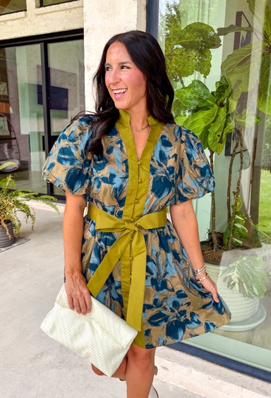 Bring timeless charm to your fall wardrobe with this beautifully detailed floral dress. Designed in a rich olive green with a stunning blue and navy floral print, this cotton dress is the perfect blend of classic and feminine. The flattering v-neckline, button-front design, and self-tie waist create a silhouette that's effortlessly polished, while the gathered back yoke adds a soft, flowy fit you'll love.