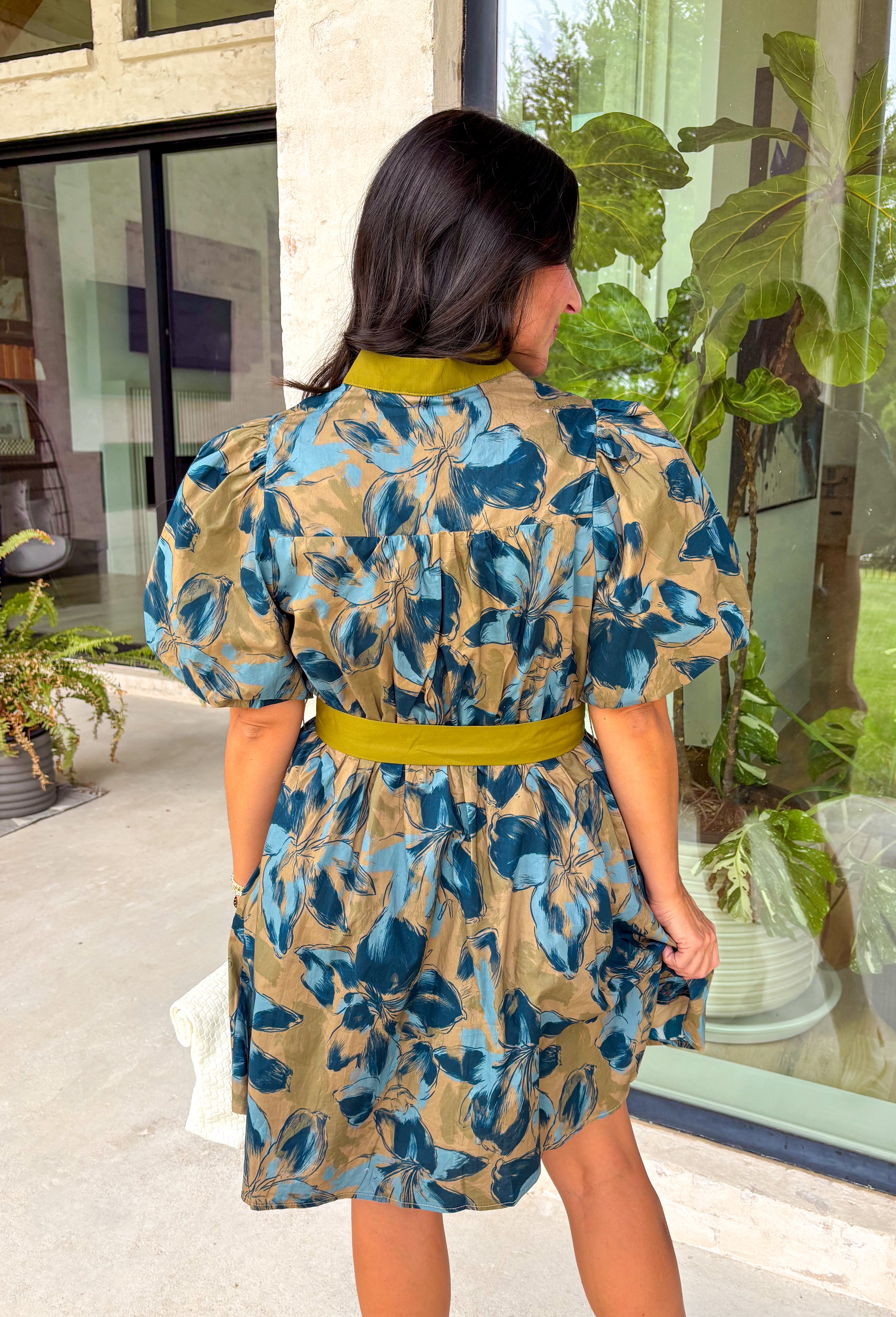 Bring timeless charm to your fall wardrobe with this beautifully detailed floral dress. Designed in a rich olive green with a stunning blue and navy floral print, this cotton dress is the perfect blend of classic and feminine. The flattering v-neckline, button-front design, and self-tie waist create a silhouette that's effortlessly polished, while the gathered back yoke adds a soft, flowy fit you'll love.