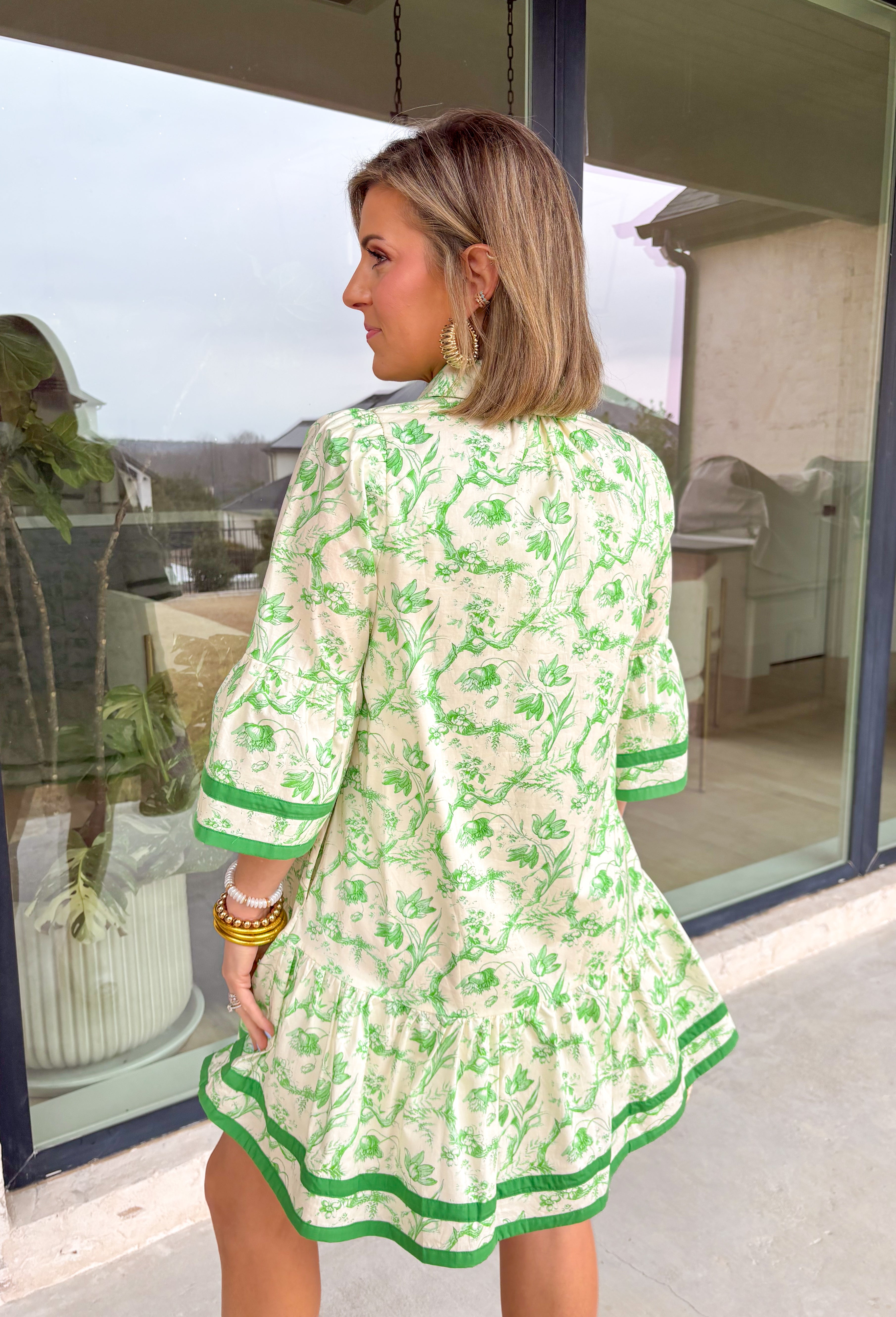 Fresh, timeless, and perfect for spring, this off-white dress by Entro is an easy favorite for sunny days ahead. Featuring a light green floral print throughout, it has a soft, feminine look that feels effortlessly wearable. Dark green trim along the sleeves and hem adds a polished contrast, while the front button details give it a classic touch. Finished with side pockets and a full lining, this dress is comfortable, flattering, and ready for everything from brunch to spring events.