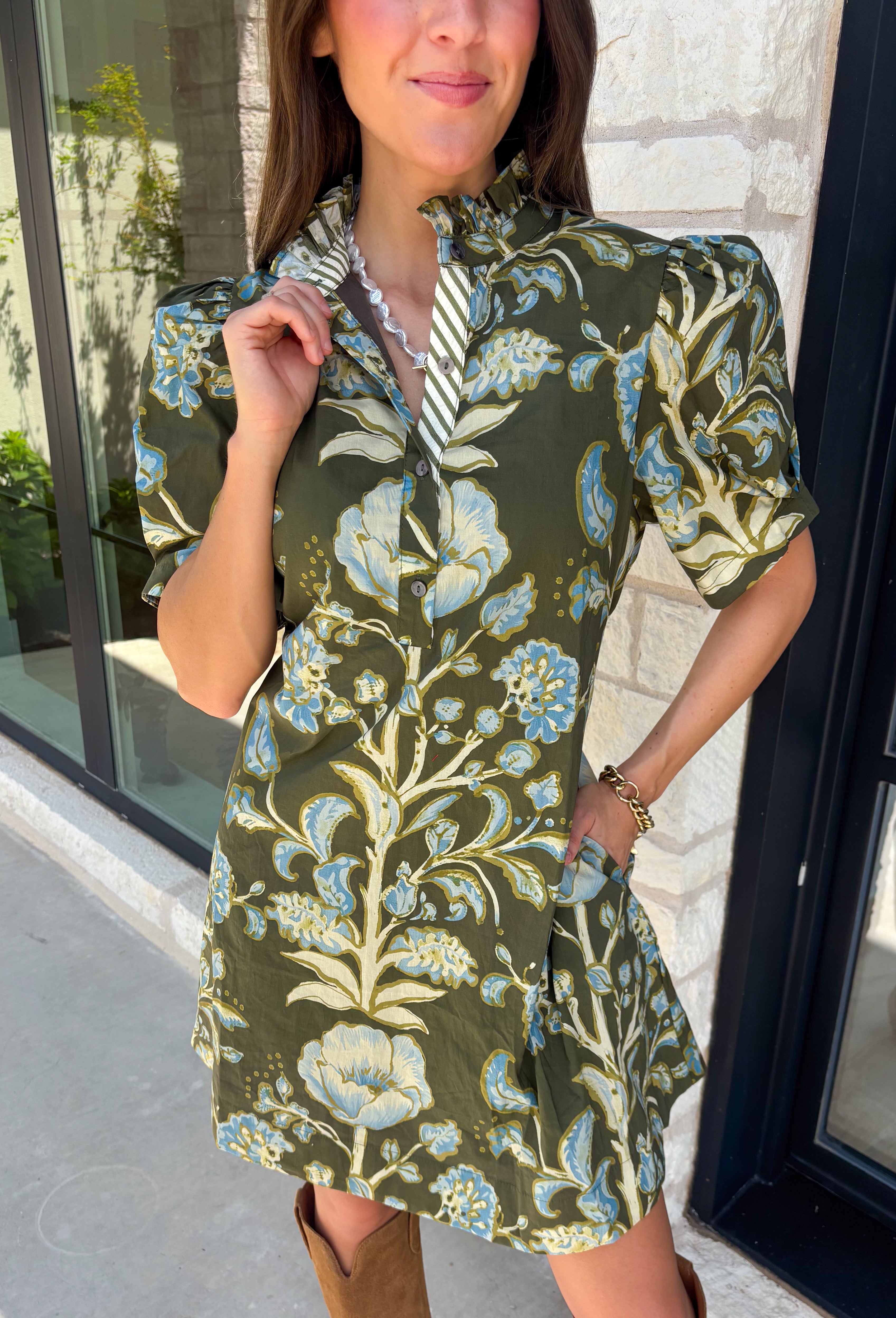 Welcome the season in effortless style with this charming floral dress! Featuring a rich olive backdrop with beautiful blue and soft green florals, this dress captures everything we love about fall. The ruffled high neckline and button front add timeless charm, while the self-tie waist in the back creates a flattering silhouette that's easy to customize. Finished with short puff sleeves, side pockets, and a fully lined design.