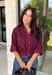 Every wardrobe needs a timeless wine blouse, and this versatile top from Entro is one you'll reach for again and again. Designed with a flattering v-neckline and button-front closure, this effortlessly polished piece transitions seamlessly from workdays to weekends. The ¾ roll-tab sleeves offer versatile styling, while the elasticized smocked back hem creates a comfortable fit with a modern touch. 