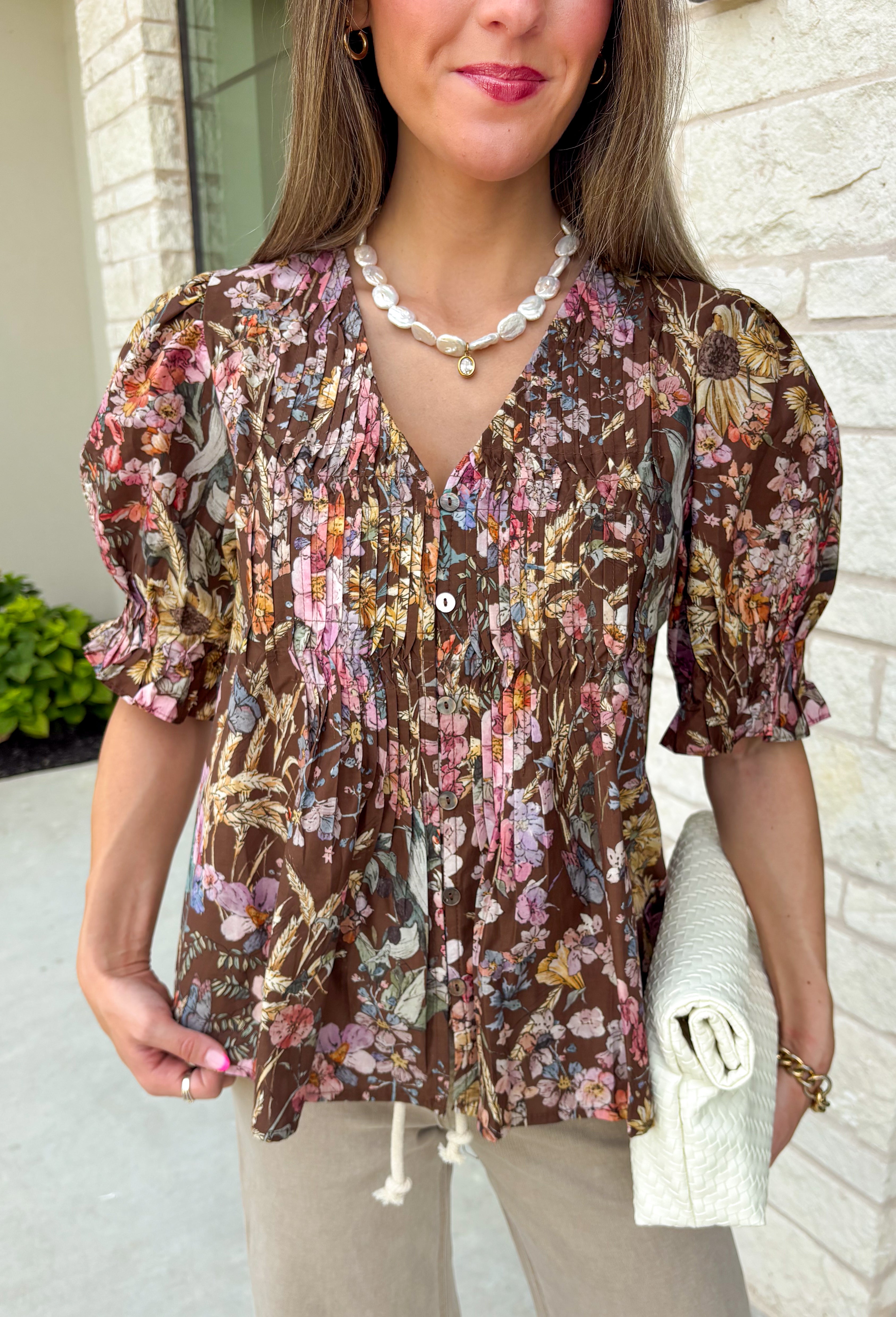 Bring a touch of timeless charm to your fall wardrobe with this beautifully detailed floral blouse. Designed in a warm brown hue with a soft neutral floral print, this top is the perfect blend of feminine and effortless. Delicate pintuck detailing, a flattering v-neckline, and button-front accents create a polished look, while the puff sleeves with smocked ruffle cuffs add just the right amount of romantic flair. 