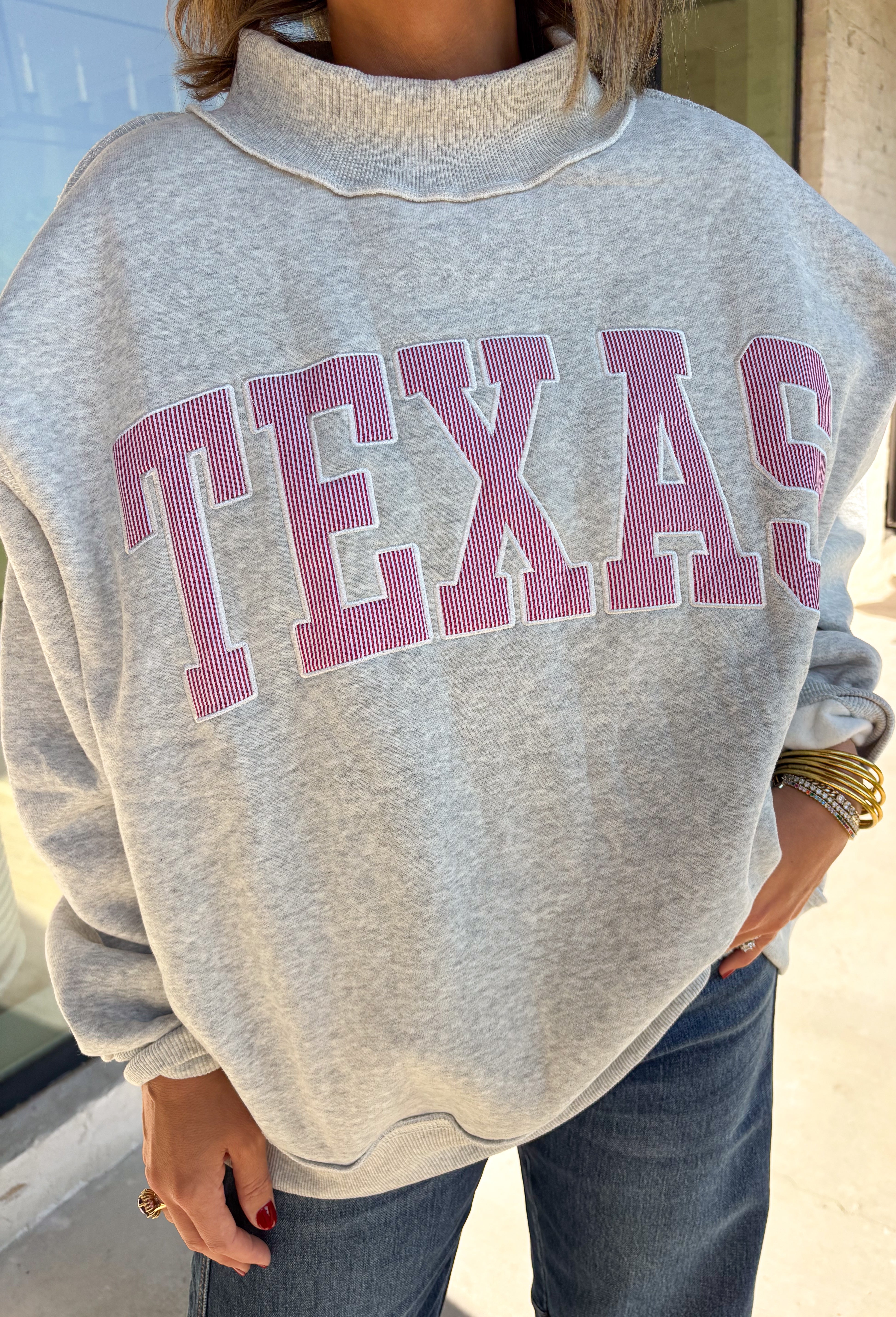 Show a little Lone Star love while staying extra cozy in this heather grey “Texas” pullover from Bucket List. The bold lettering features a crimson and white stripe print, with each letter outlined in white embroidery for added texture and a standout finish. A mock neckline and long sleeves give it a cozy cold-weather feel, while the fleece-lined interior makes it perfect for crisp fall mornings and chilly weekends. 