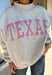 Show a little Lone Star love while staying extra cozy in this heather grey “Texas” pullover from Bucket List. The bold lettering features a crimson and white stripe print, with each letter outlined in white embroidery for added texture and a standout finish. A mock neckline and long sleeves give it a cozy cold-weather feel, while the fleece-lined interior makes it perfect for crisp fall mornings and chilly weekends. 