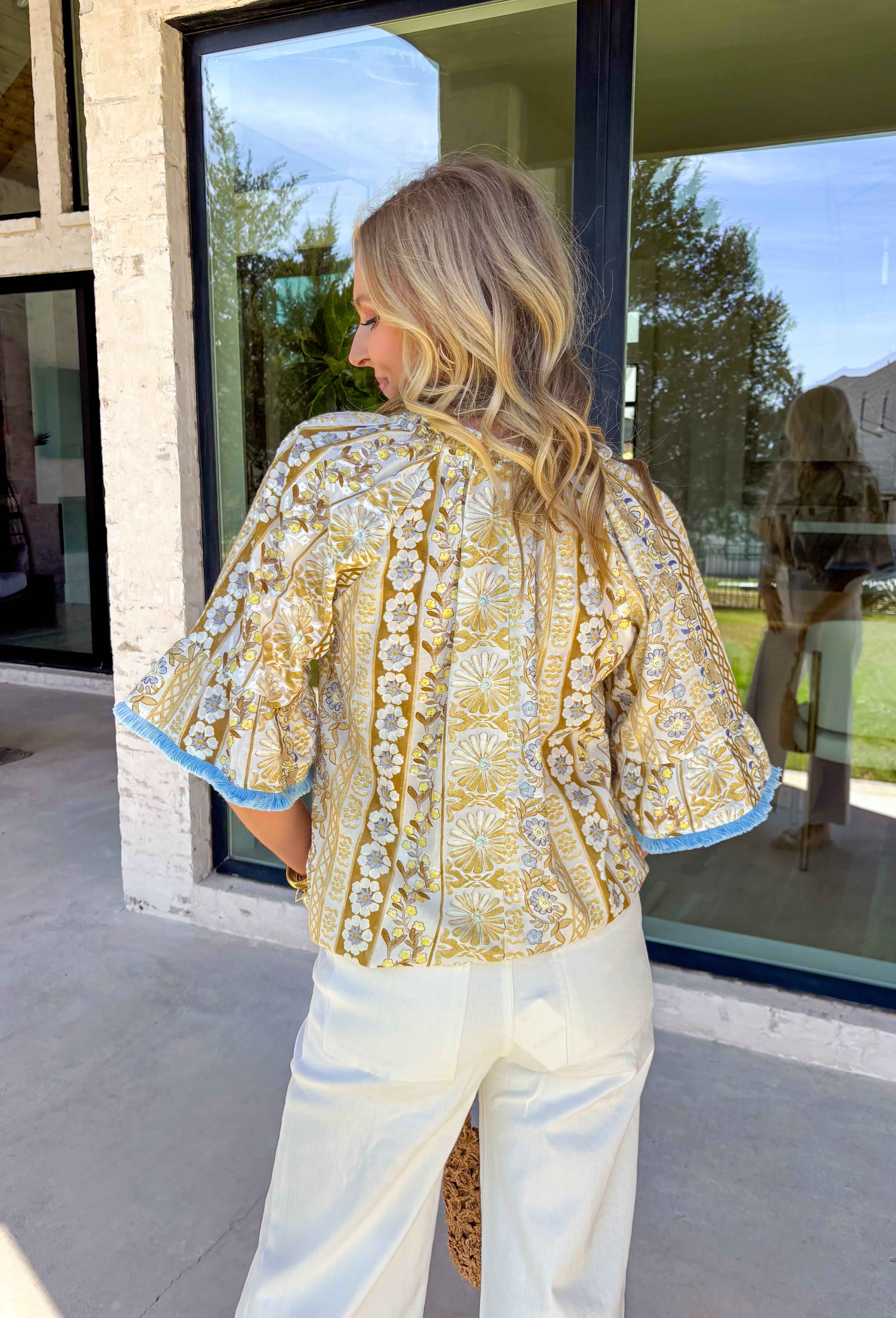 This sweet little spring top is basically sunshine in outfit form! Featuring a crisp white base with soft yellow florals and the prettiest hints of baby blue, it’s the kind of piece that instantly brightens your day. The subtle ruffle neckline keeps it feminine and fun, while the front tie with tassel details adds that effortless “threw it on and still look cute” vibe. 