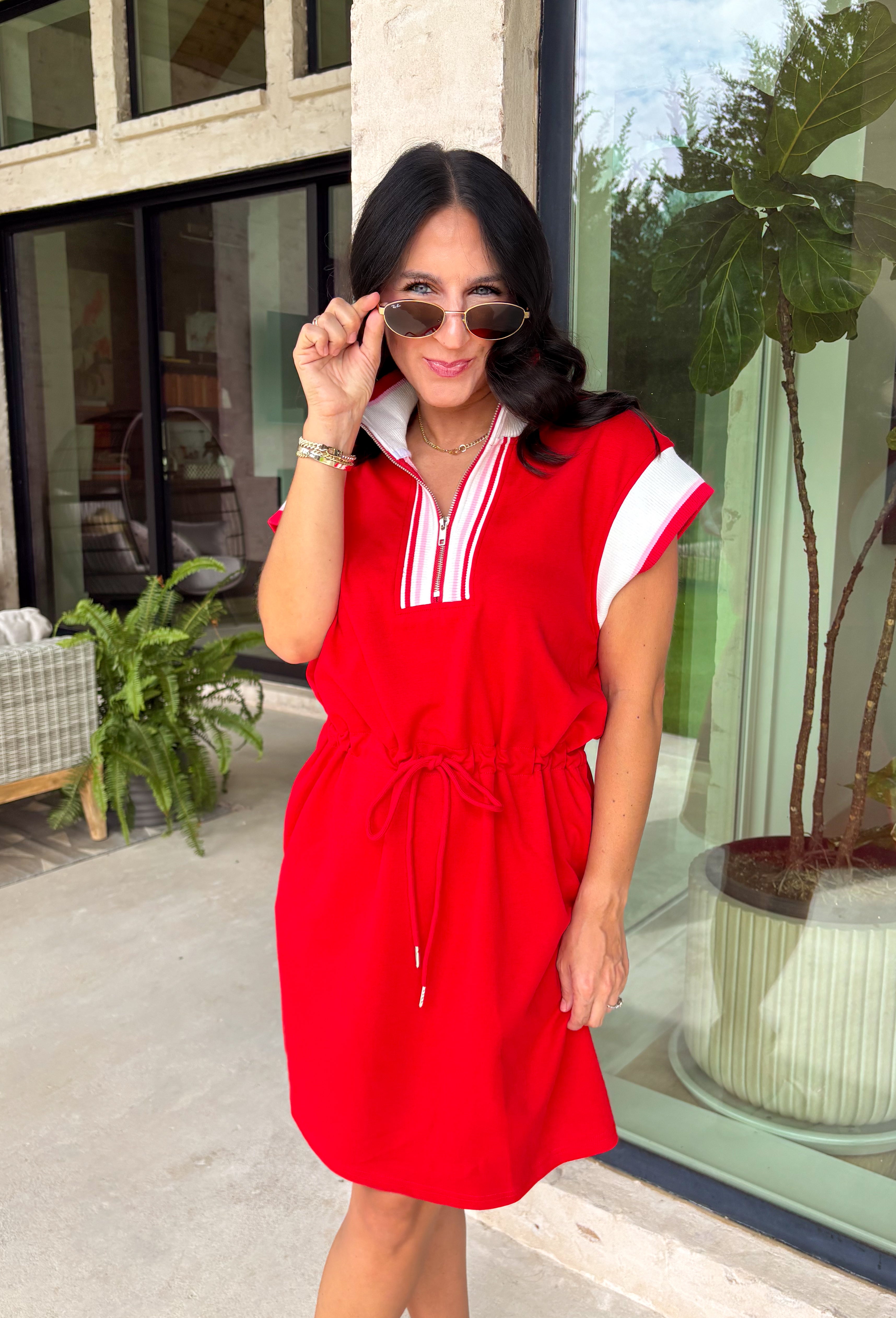 This red short sleeve dress is the perfect mix of sporty and cute for summer! Designed with a quarter zip neckline and elastic self-tie waist, it gives you that flattering, adjustable fit while keeping things comfortable for all-day wear. The red, pink, and white athletic stripe detail along the front, neckline, and cuffs adds a fun, energetic touch that makes this one stand out. Finished with side pockets (always a win), it’s perfect for everything from errands to casual plans.