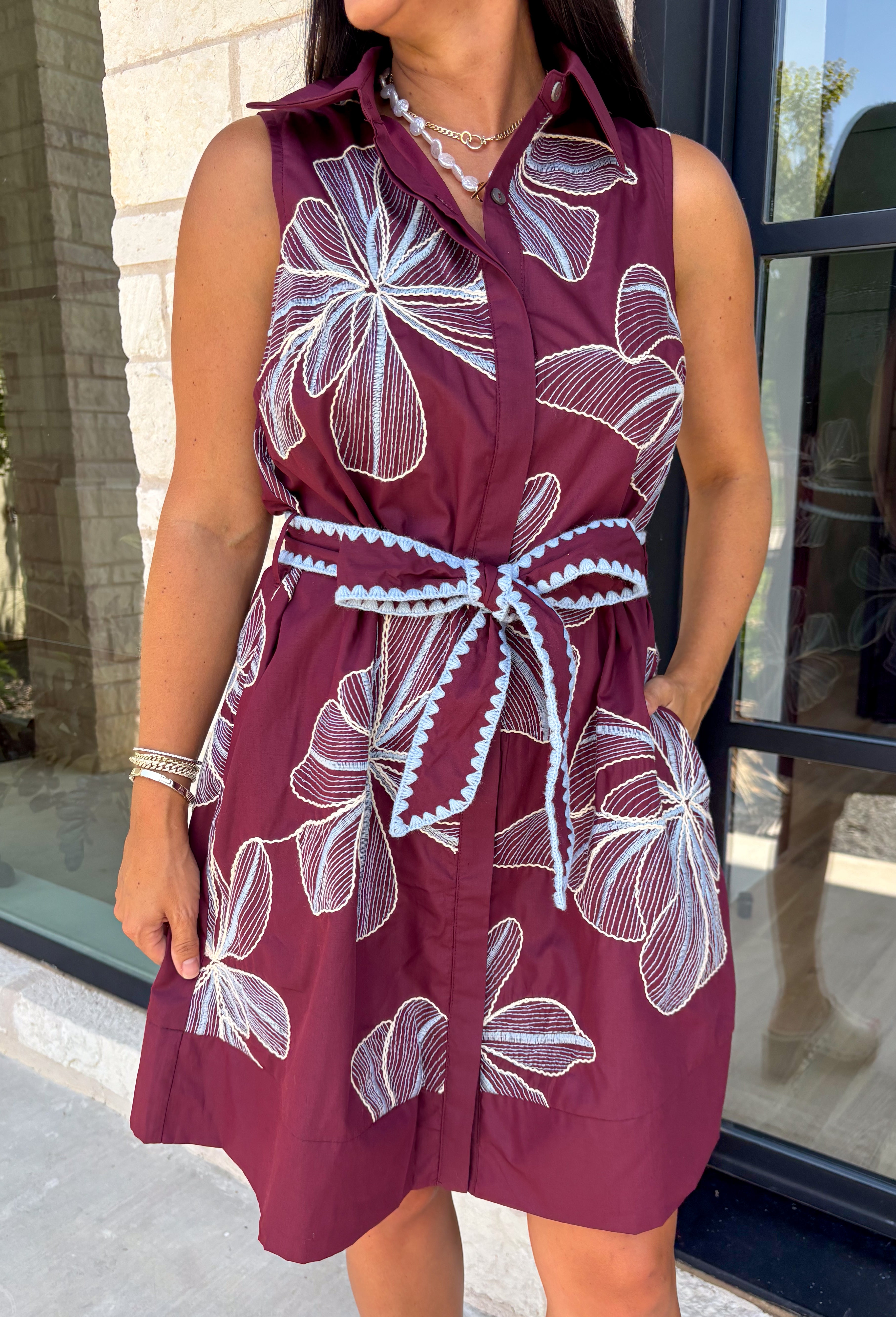 Rich fall colors and beautiful embroidered details make this dress an instant seasonal favorite! Crafted in a gorgeous wine hue, this sleeveless dress features delicate light blue and off white floral embroidery that creates a charming, handcrafted look. The button front and self-tie waist belt with playful contrast pom trim create a flattering silhouette, while side pockets and a fully lined design make it as practical as it is beautiful. 