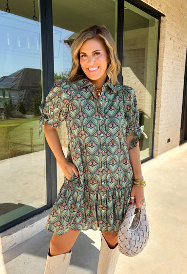 Embrace the beauty of the season in this charming dress from Entro. Featuring a gorgeous floral print in shades of hunter green, muted green, peach, and brown, this feminine dress is perfect for everything from weekend brunches to seasonal celebrations. The button-front design and flattering neckline create a timeless silhouette, while the ¾ puff sleeves with delicate tie details add a playful touch. 
