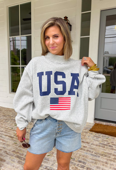 This grey reversible pullover is the ultimate cozy-meets-festive piece for your summer celebrations! Designed with two looks in one, one side features “USA” in navy with a flag design, while the other side is fleece lined with “America” in blue for a softer, cozier feel. The mock neck adds an elevated touch, making it perfect for those cooler summer nights—think fireworks, lake evenings, or early morning coffee runs. It’s an easy throw-on layer that still feels fun and patriotic. 