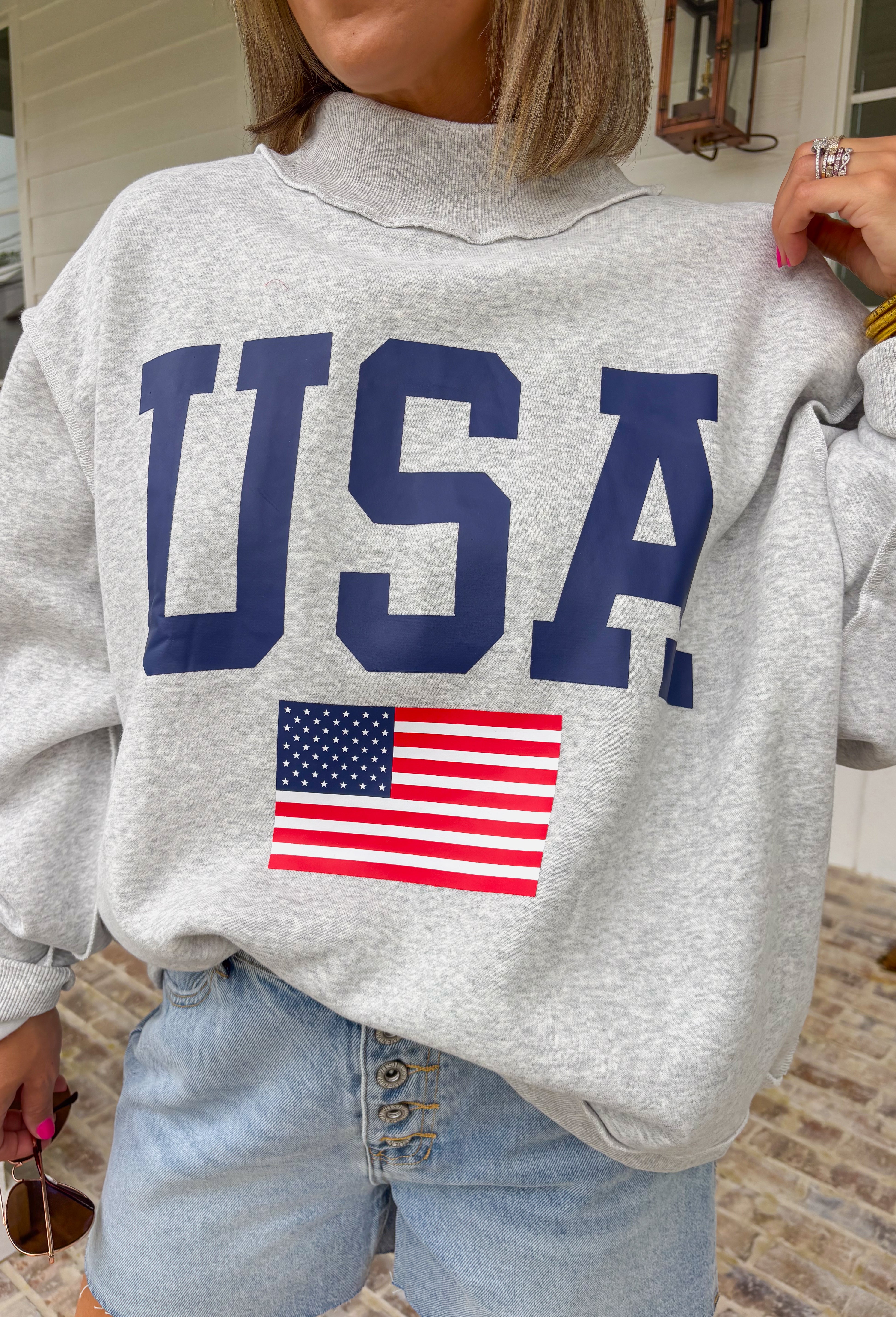This grey reversible pullover is the ultimate cozy-meets-festive piece for your summer celebrations! Designed with two looks in one, one side features “USA” in navy with a flag design, while the other side is fleece lined with “America” in blue for a softer, cozier feel. The mock neck adds an elevated touch, making it perfect for those cooler summer nights—think fireworks, lake evenings, or early morning coffee runs. It’s an easy throw-on layer that still feels fun and patriotic. 