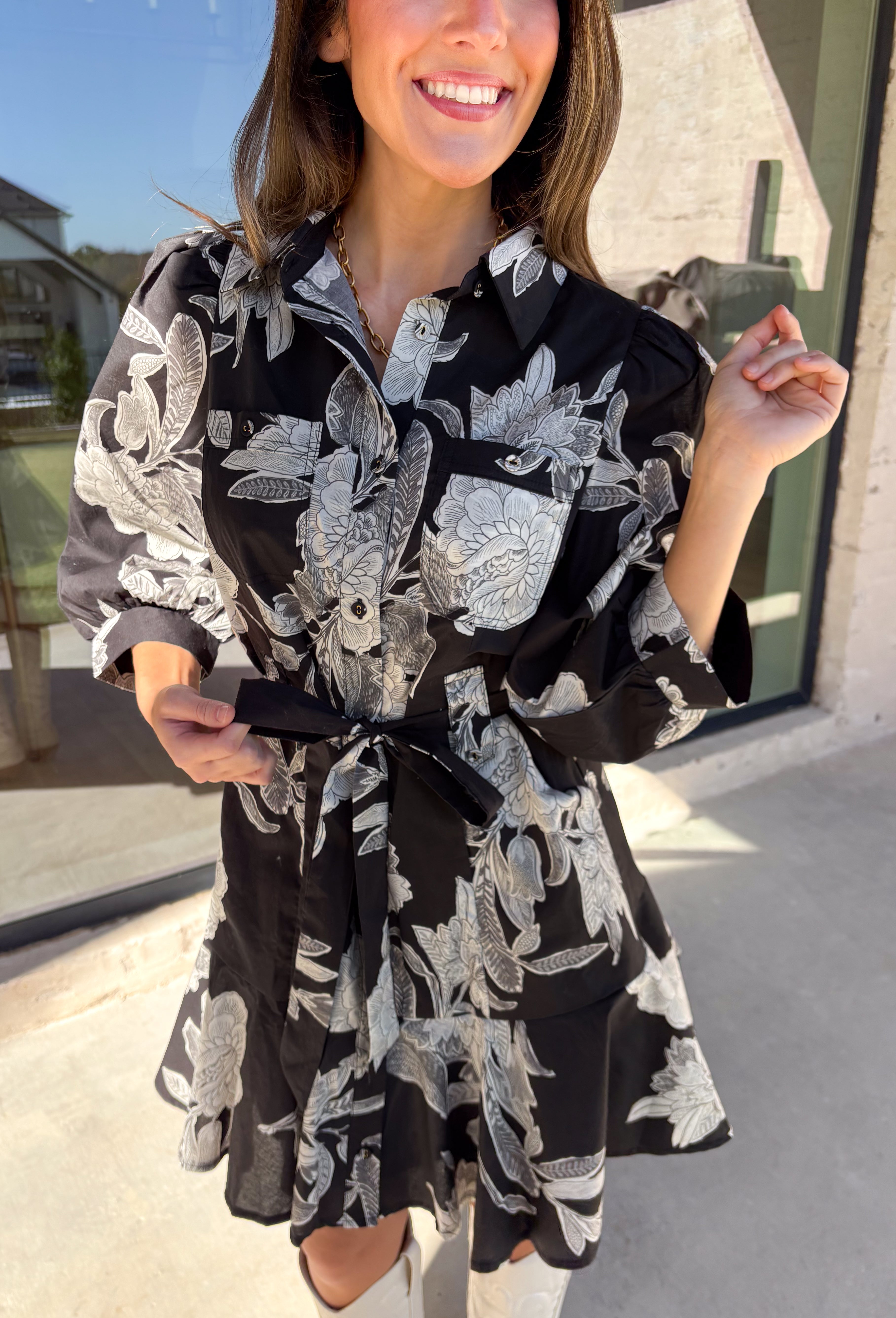 This black floral dress by Entro is a timeless favorite with a touch of charm. Designed with an off-white and gray floral print, it features ¾ puff sleeves, a button-front closure, and a flattering self-tie waist belt. The chest pocket adds a subtle utility touch, while the tiered ruffle hem and side pockets keep the look feminine and functional. This dress is perfect for any special occasion!