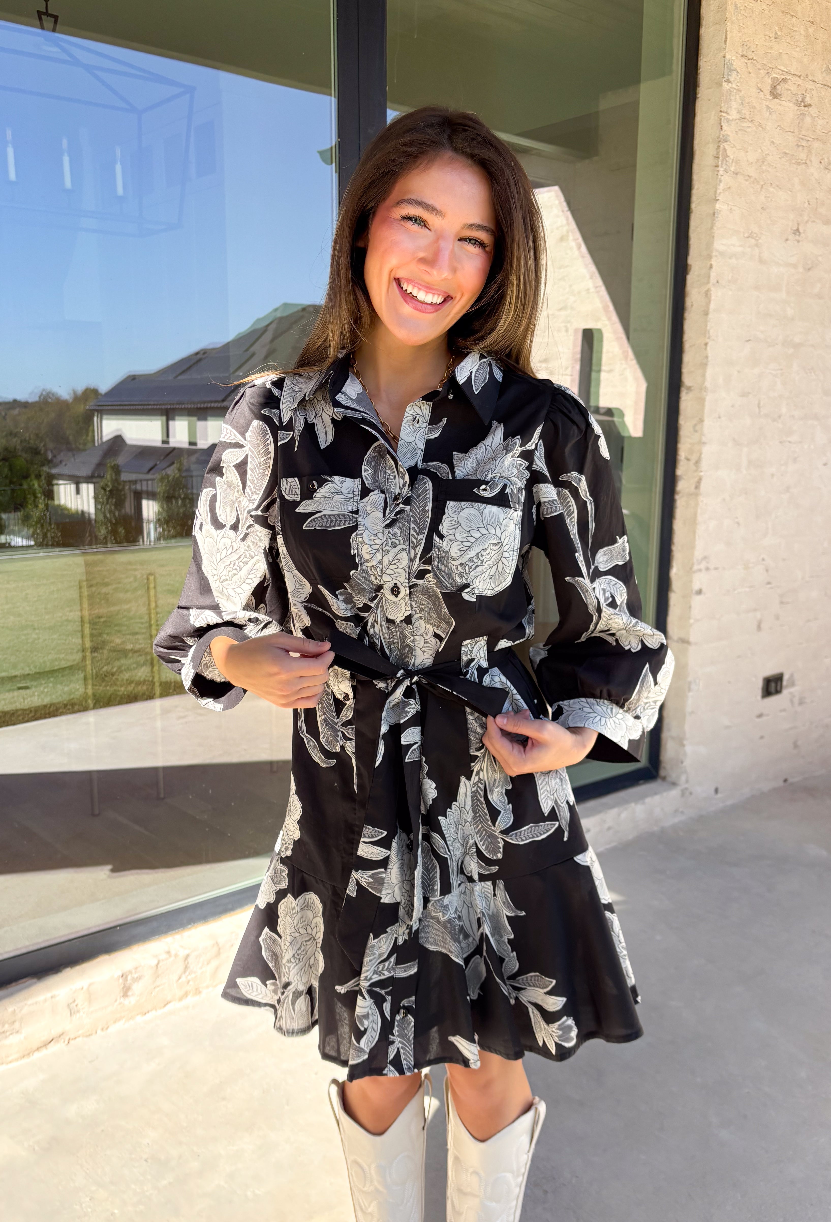 This black floral dress by Entro is a timeless favorite with a touch of charm. Designed with an off-white and gray floral print, it features ¾ puff sleeves, a button-front closure, and a flattering self-tie waist belt. The chest pocket adds a subtle utility touch, while the tiered ruffle hem and side pockets keep the look feminine and functional. This dress is perfect for any special occasion!