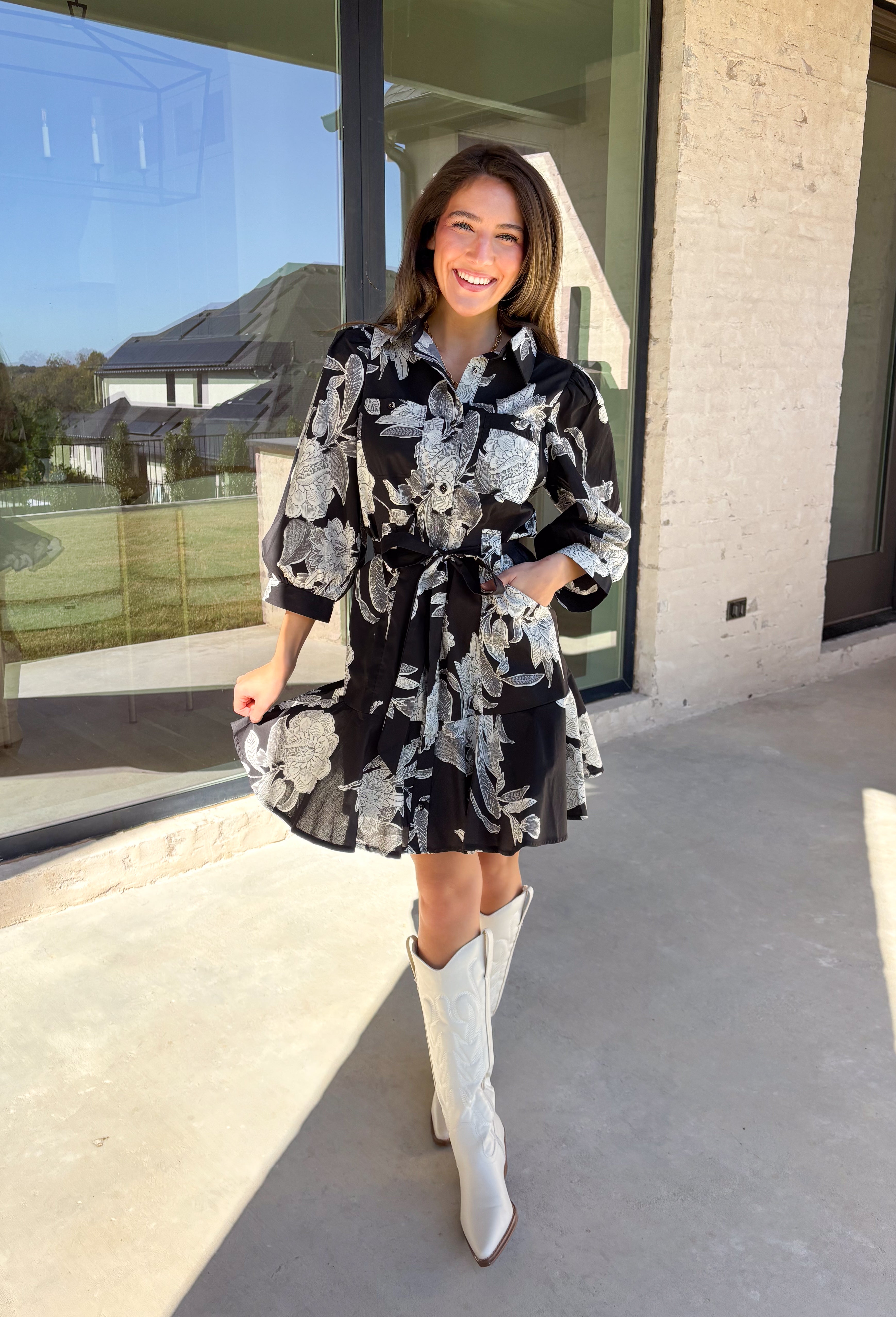 This black floral dress by Entro is a timeless favorite with a touch of charm. Designed with an off-white and gray floral print, it features ¾ puff sleeves, a button-front closure, and a flattering self-tie waist belt. The chest pocket adds a subtle utility touch, while the tiered ruffle hem and side pockets keep the look feminine and functional. This dress is perfect for any special occasion!