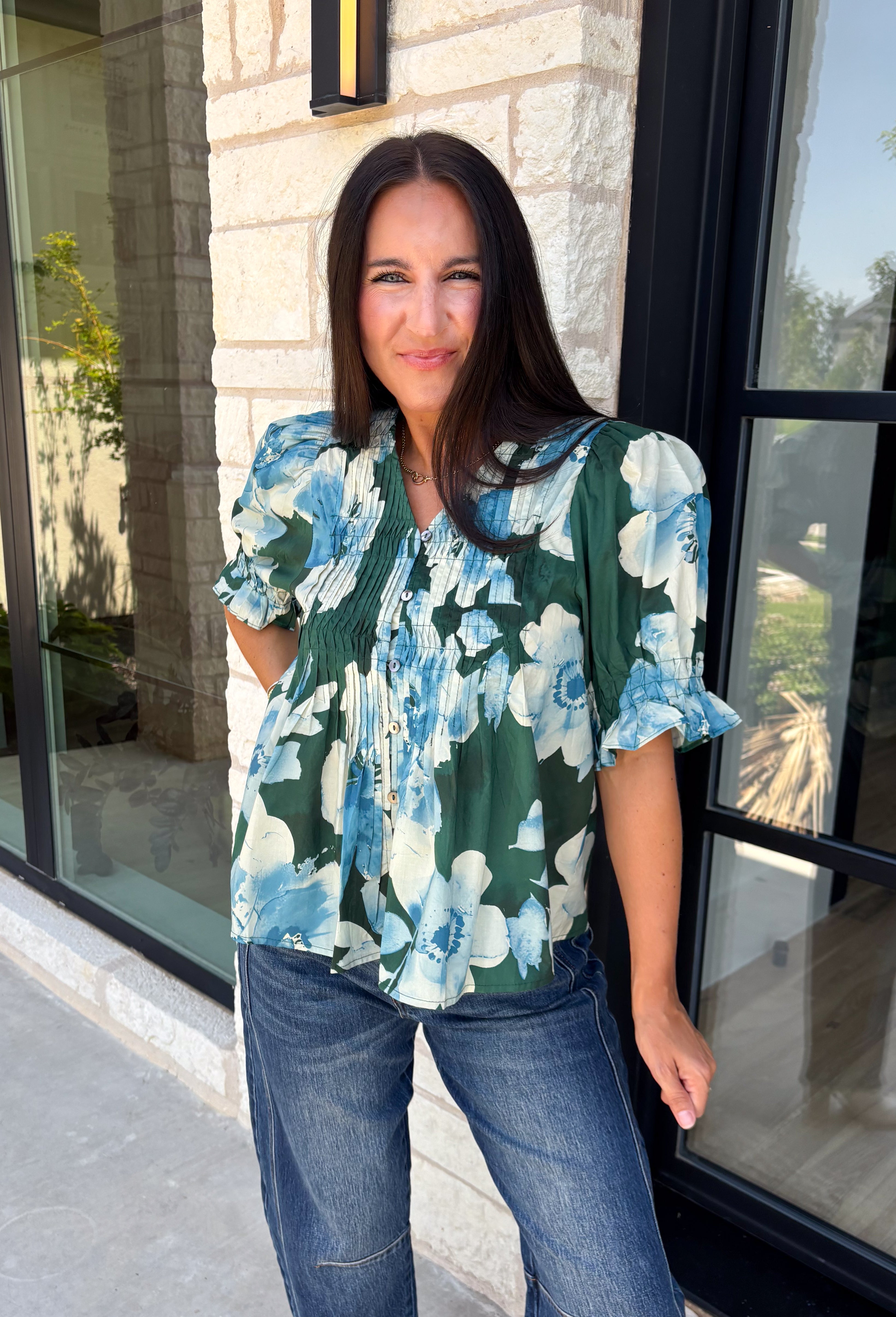Bring a fresh take on fall florals to your wardrobe with this beautiful hunter green top. Featuring a delicate blue and off white floral print, this timeless top is perfect for everything from office days to weekend outings. The pintuck detailing along the front adds an elevated touch, while the button front and short puff sleeves create a feminine silhouette you'll love season after season. 
