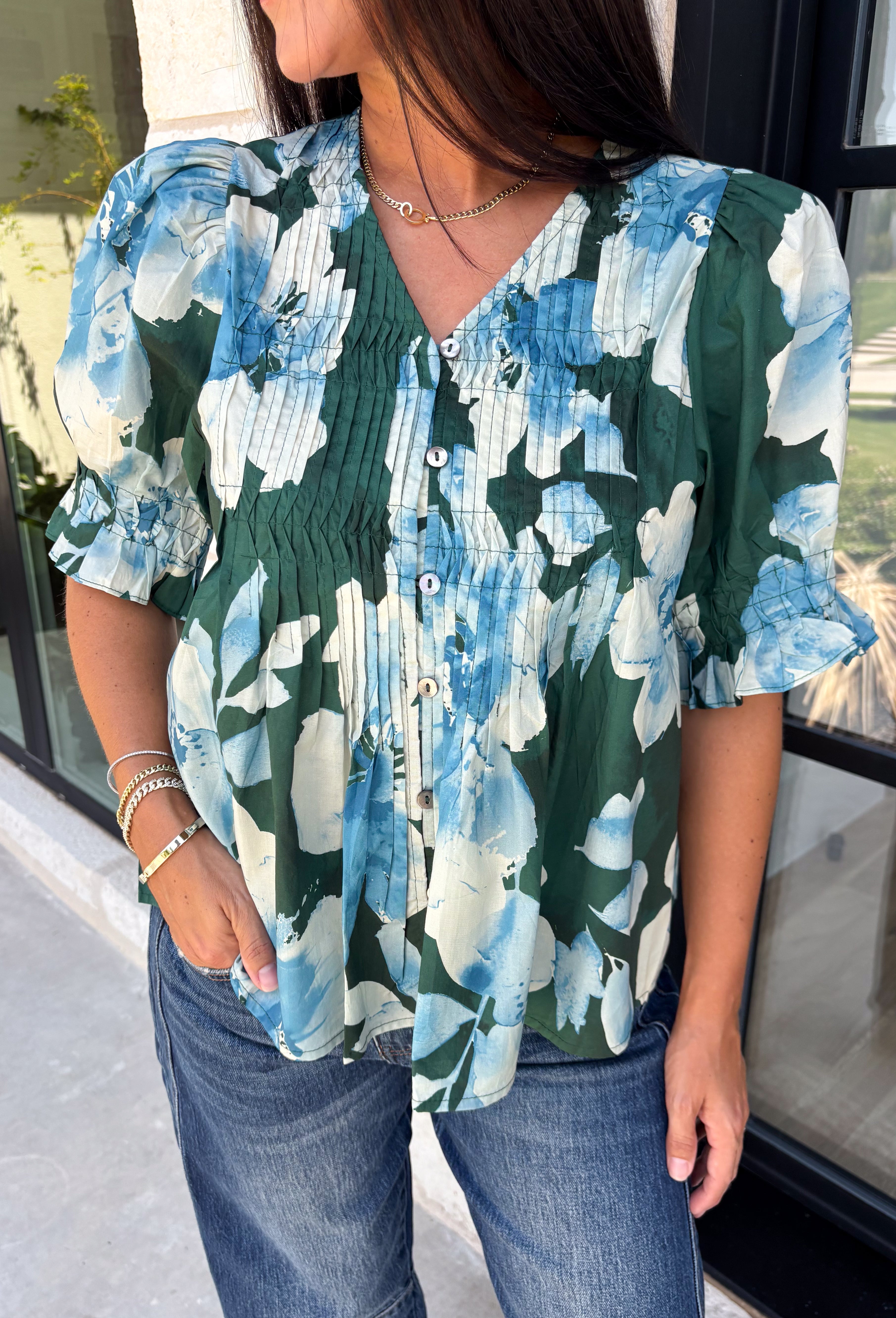 Bring a fresh take on fall florals to your wardrobe with this beautiful hunter green top. Featuring a delicate blue and off white floral print, this timeless top is perfect for everything from office days to weekend outings. The pintuck detailing along the front adds an elevated touch, while the button front and short puff sleeves create a feminine silhouette you'll love season after season. 