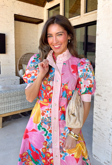 Bright, joyful, and bursting with spring color, this white short sleeve button-down dress by Entro is a total showstopper. Featuring a bold floral print in pink, red, blue, yellow, green, and orange, it brings instant happy vibes to your wardrobe. The ruffle neckline is trimmed with pink and white stripes for a fun pop of contrast, with matching stripe detail running down the front and along the sleeve cuffs. Smocking at the back of the sleeves adds comfort and shape, while side pockets and a lining make it