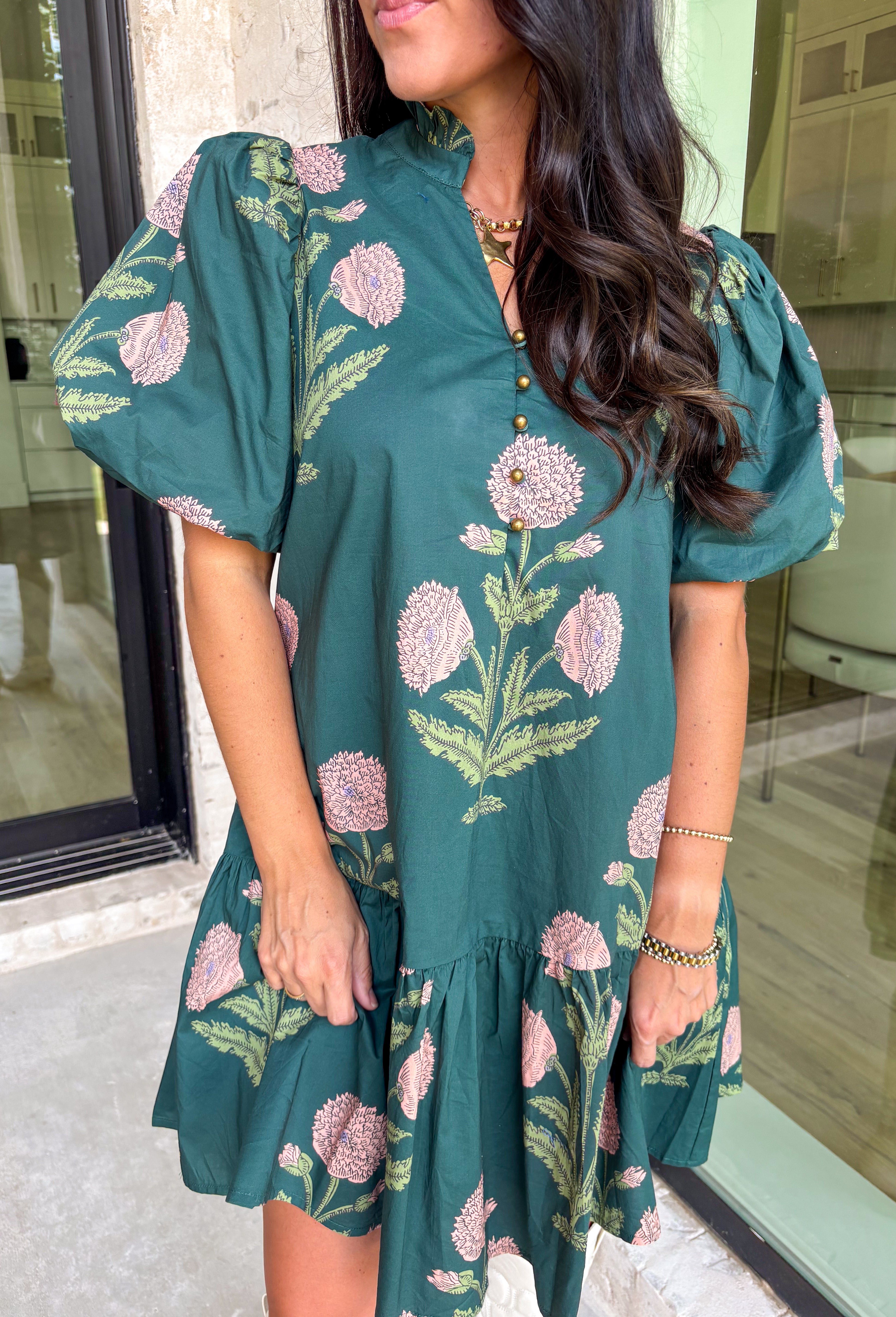 Rich, romantic, and made for fall days, this hunter green mini dress is covered in a warm seasonal floral print. The ruffle neck band collar with gold accent button closure adds a polished touch, while the short bubble sleeves and flouncy ruffle hem keep it playful and feminine. Side pockets bring in that practical detail we love, and the fully lined cotton design makes it comfortable from day to night. Perfect for everything from brunch to seasonal gatherings.