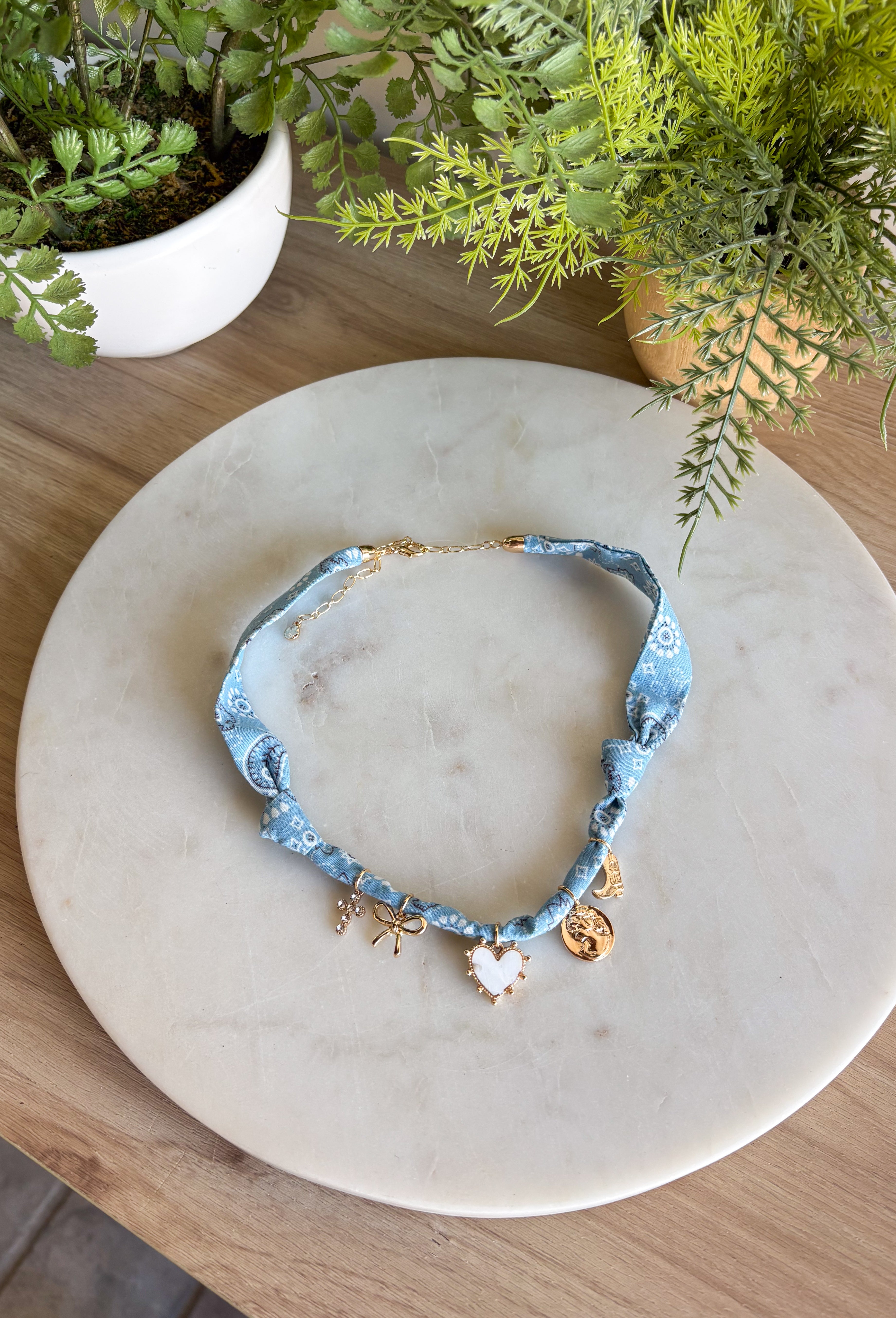 Playful and trendy, this light blue bandana necklace is full of personality. Detailed with a mix of eclectic charms—including a sparkling crystal cross, gold bow, white heart, gold pendant, and a gold cowboy boot—it’s the perfect statement piece to add fun flair to any outfit. With a 15" length and 3" extender, you can adjust it to layer with other necklaces or let it shine on its own. A must-have for adding a pop of charm to your wardrobe.