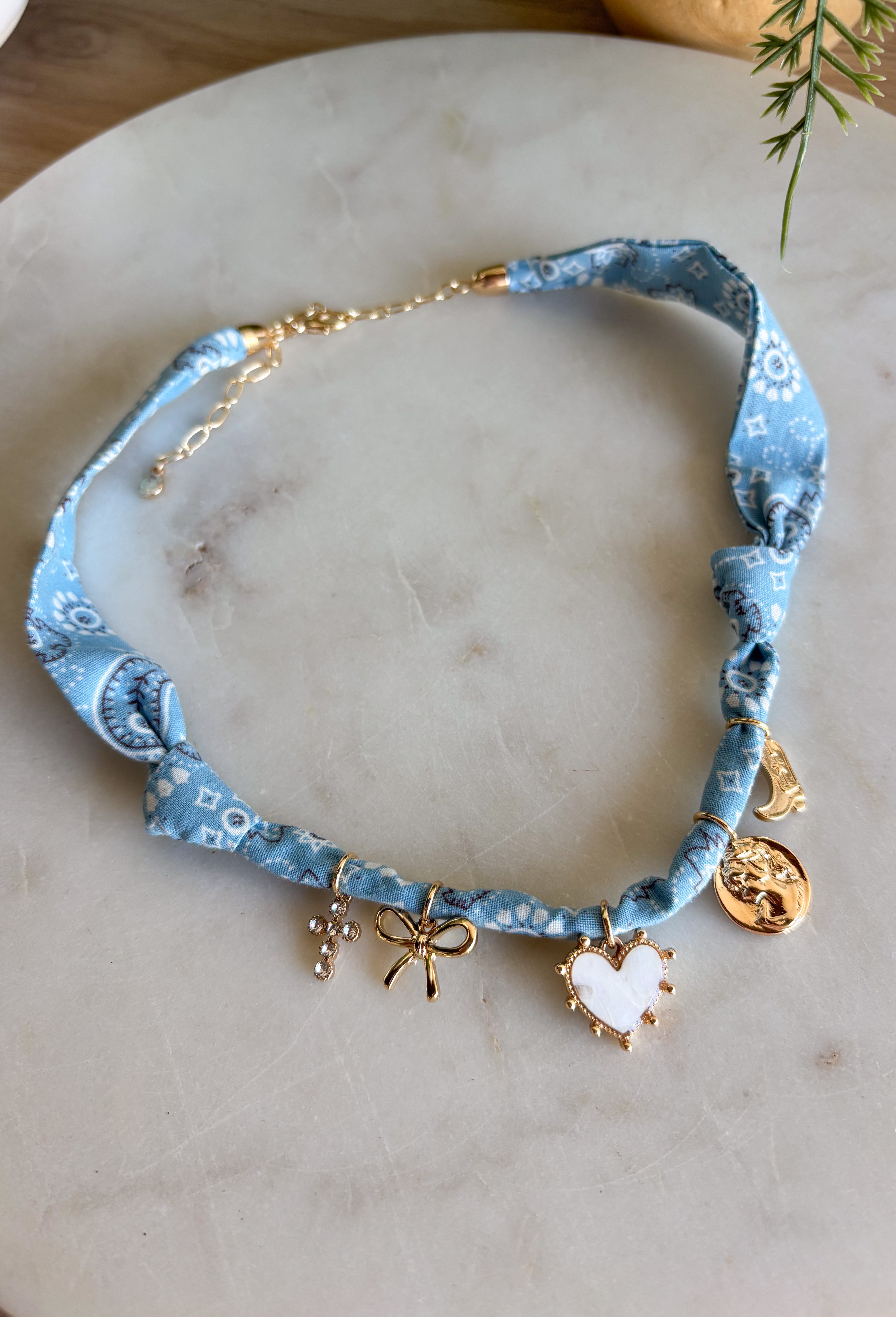 Playful and trendy, this light blue bandana necklace is full of personality. Detailed with a mix of eclectic charms—including a sparkling crystal cross, gold bow, white heart, gold pendant, and a gold cowboy boot—it’s the perfect statement piece to add fun flair to any outfit. With a 15" length and 3" extender, you can adjust it to layer with other necklaces or let it shine on its own. A must-have for adding a pop of charm to your wardrobe.