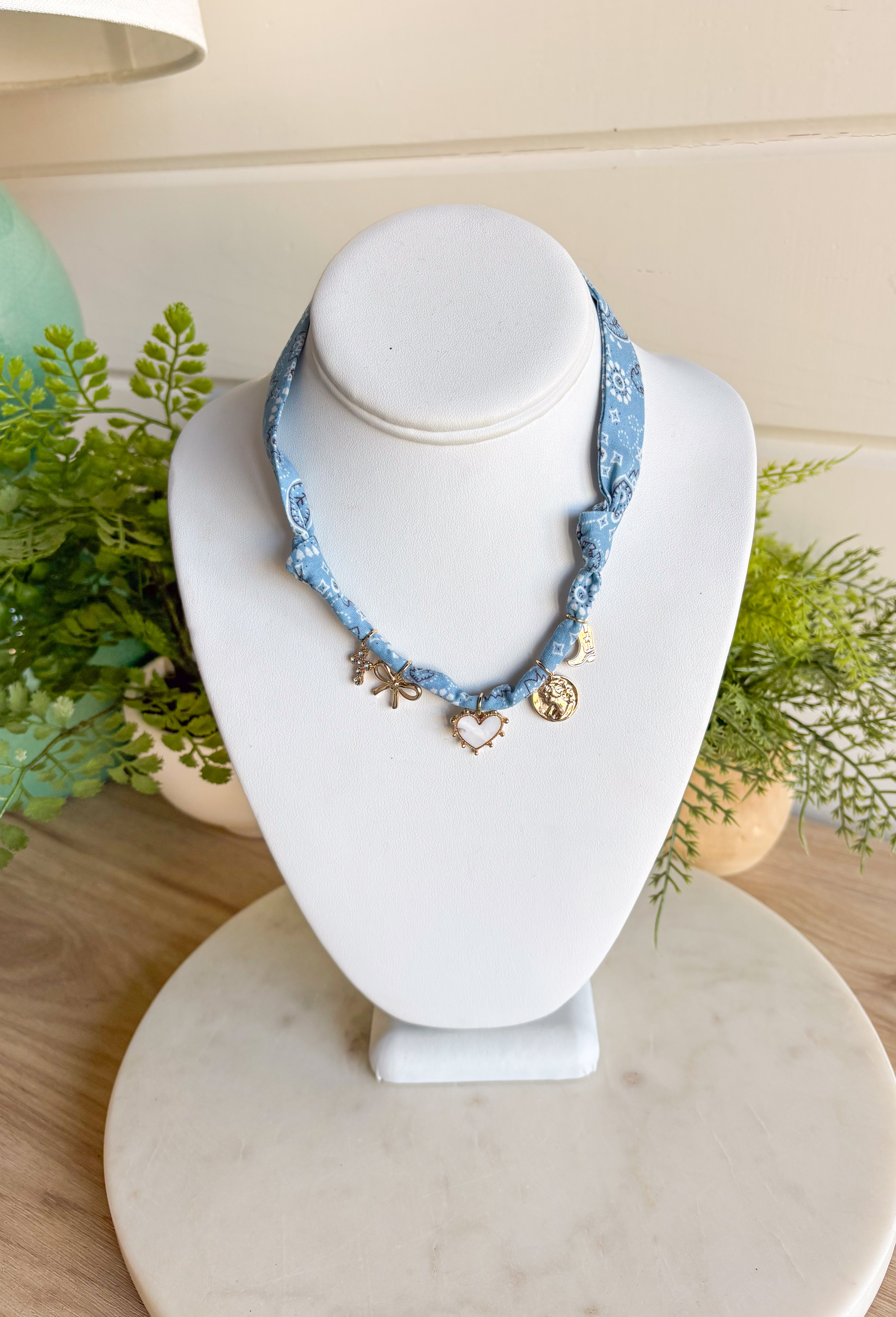 Playful and trendy, this light blue bandana necklace is full of personality. Detailed with a mix of eclectic charms—including a sparkling crystal cross, gold bow, white heart, gold pendant, and a gold cowboy boot—it’s the perfect statement piece to add fun flair to any outfit. With a 15" length and 3" extender, you can adjust it to layer with other necklaces or let it shine on its own. A must-have for adding a pop of charm to your wardrobe.