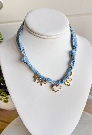 Playful and trendy, this light blue bandana necklace is full of personality. Detailed with a mix of eclectic charms—including a sparkling crystal cross, gold bow, white heart, gold pendant, and a gold cowboy boot—it’s the perfect statement piece to add fun flair to any outfit. With a 15" length and 3" extender, you can adjust it to layer with other necklaces or let it shine on its own. A must-have for adding a pop of charm to your wardrobe.