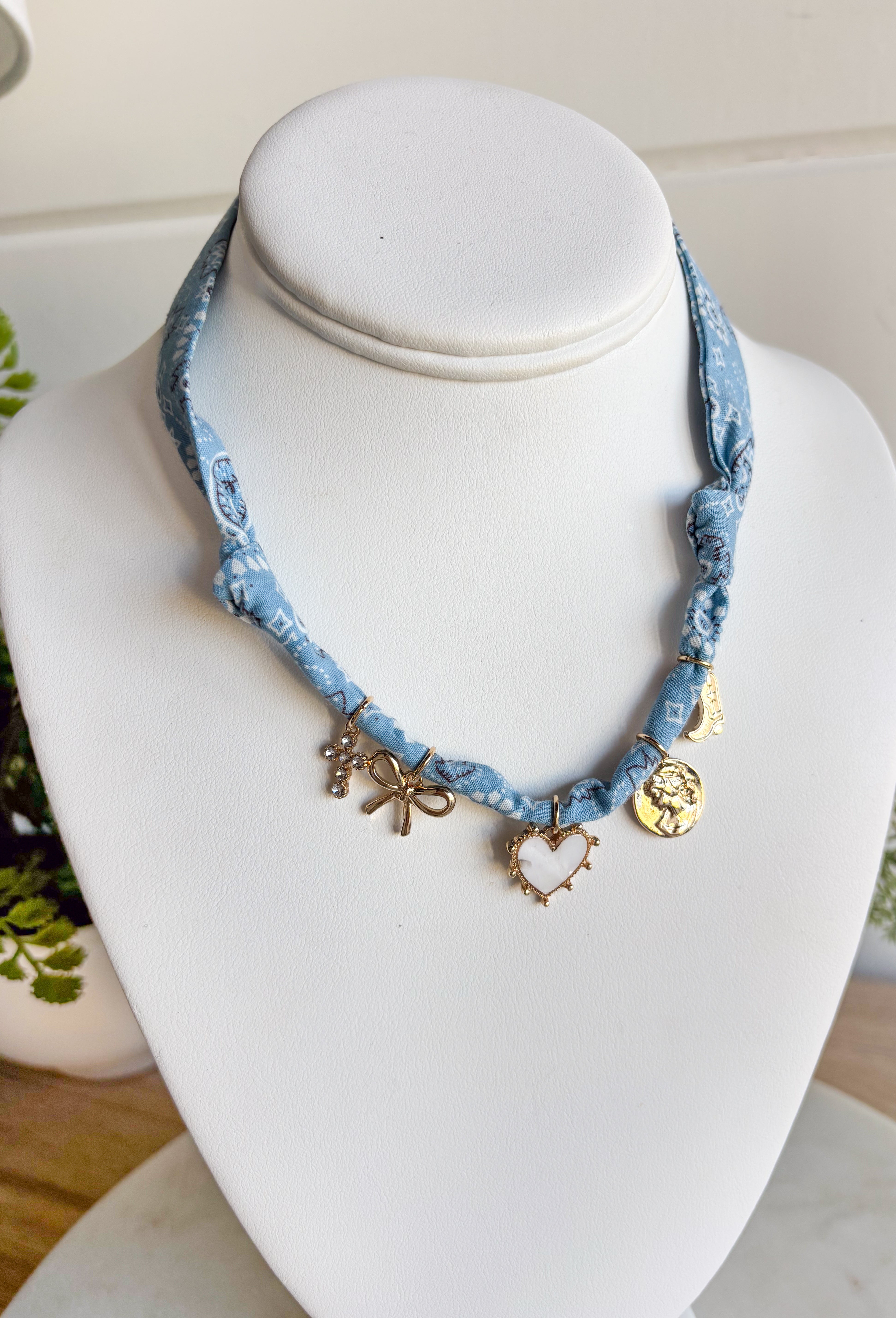 Playful and trendy, this light blue bandana necklace is full of personality. Detailed with a mix of eclectic charms—including a sparkling crystal cross, gold bow, white heart, gold pendant, and a gold cowboy boot—it’s the perfect statement piece to add fun flair to any outfit. With a 15" length and 3" extender, you can adjust it to layer with other necklaces or let it shine on its own. A must-have for adding a pop of charm to your wardrobe.
