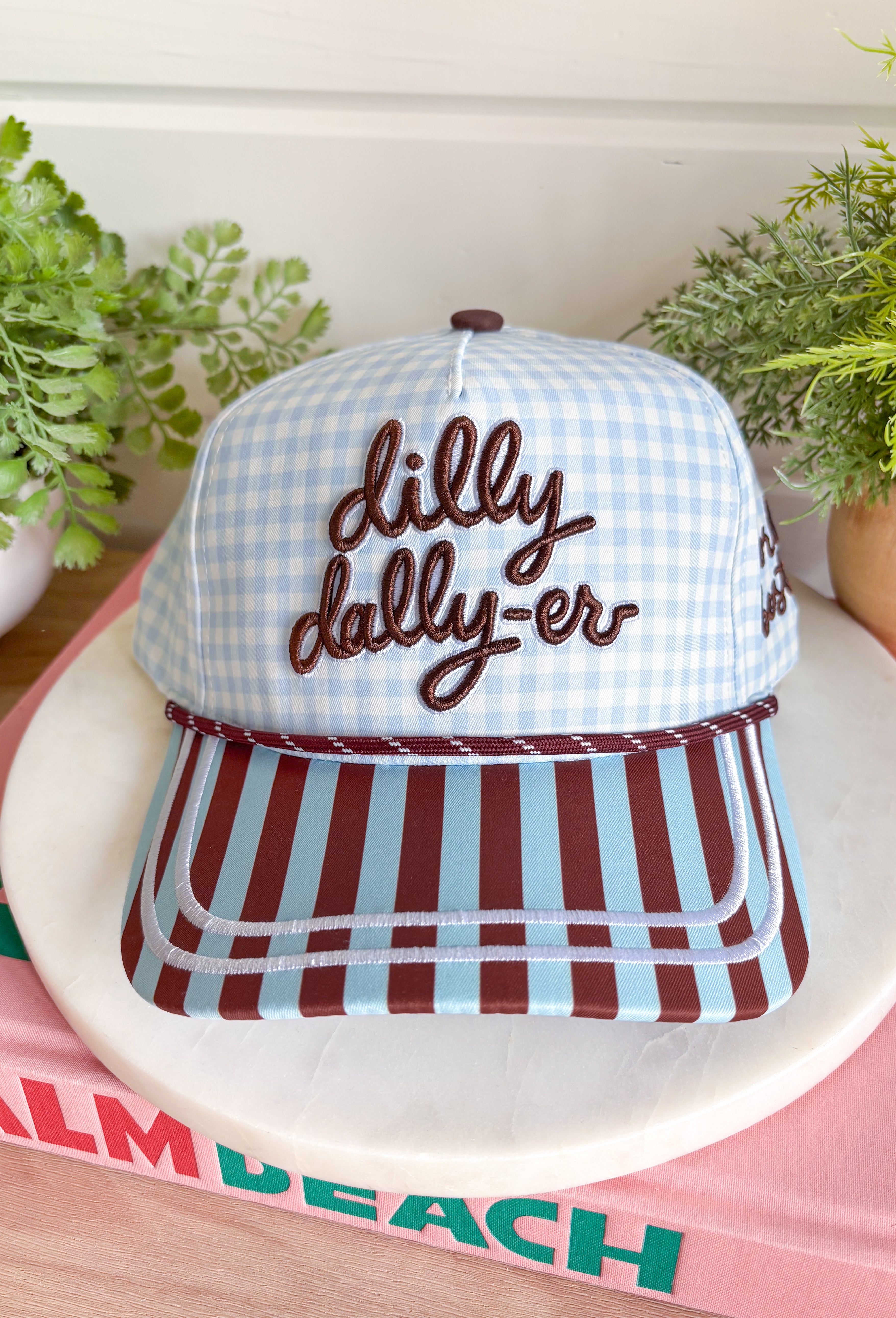 Bring a little fun to your everyday look with this charming trucker hat that's full of playful personality. Featuring a light blue and white gingham front paired with a light blue and brown striped bill, this unique hat is finished with white embroidered detailing along the bill for an extra touch of texture. The front is embroidered in chocolate brown with "Dilly Dally-er," while the side proudly reads "World's Best" with a ribbon. 