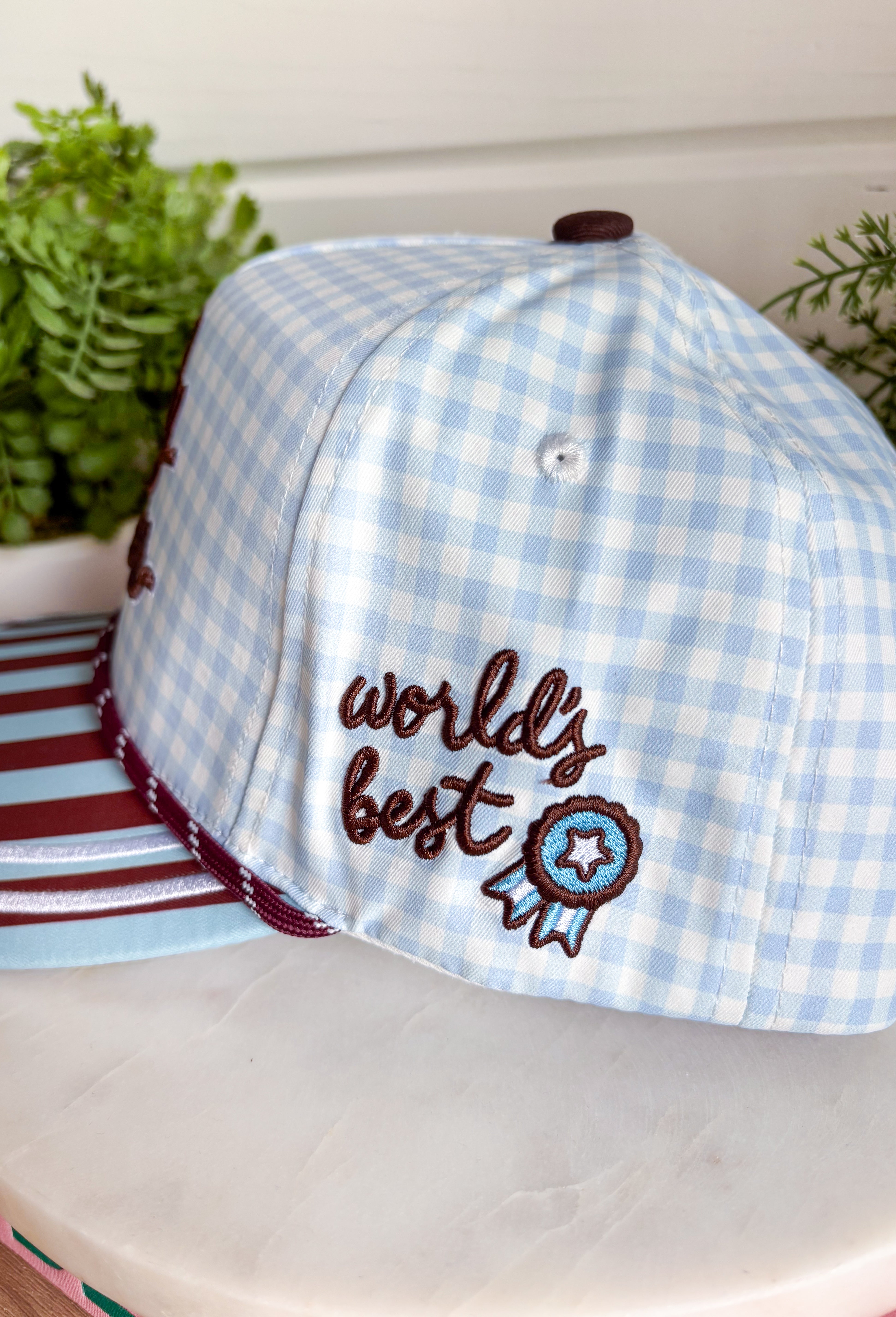 Bring a little fun to your everyday look with this charming trucker hat that's full of playful personality. Featuring a light blue and white gingham front paired with a light blue and brown striped bill, this unique hat is finished with white embroidered detailing along the bill for an extra touch of texture. The front is embroidered in chocolate brown with "Dilly Dally-er," while the side proudly reads "World's Best" with a ribbon. 