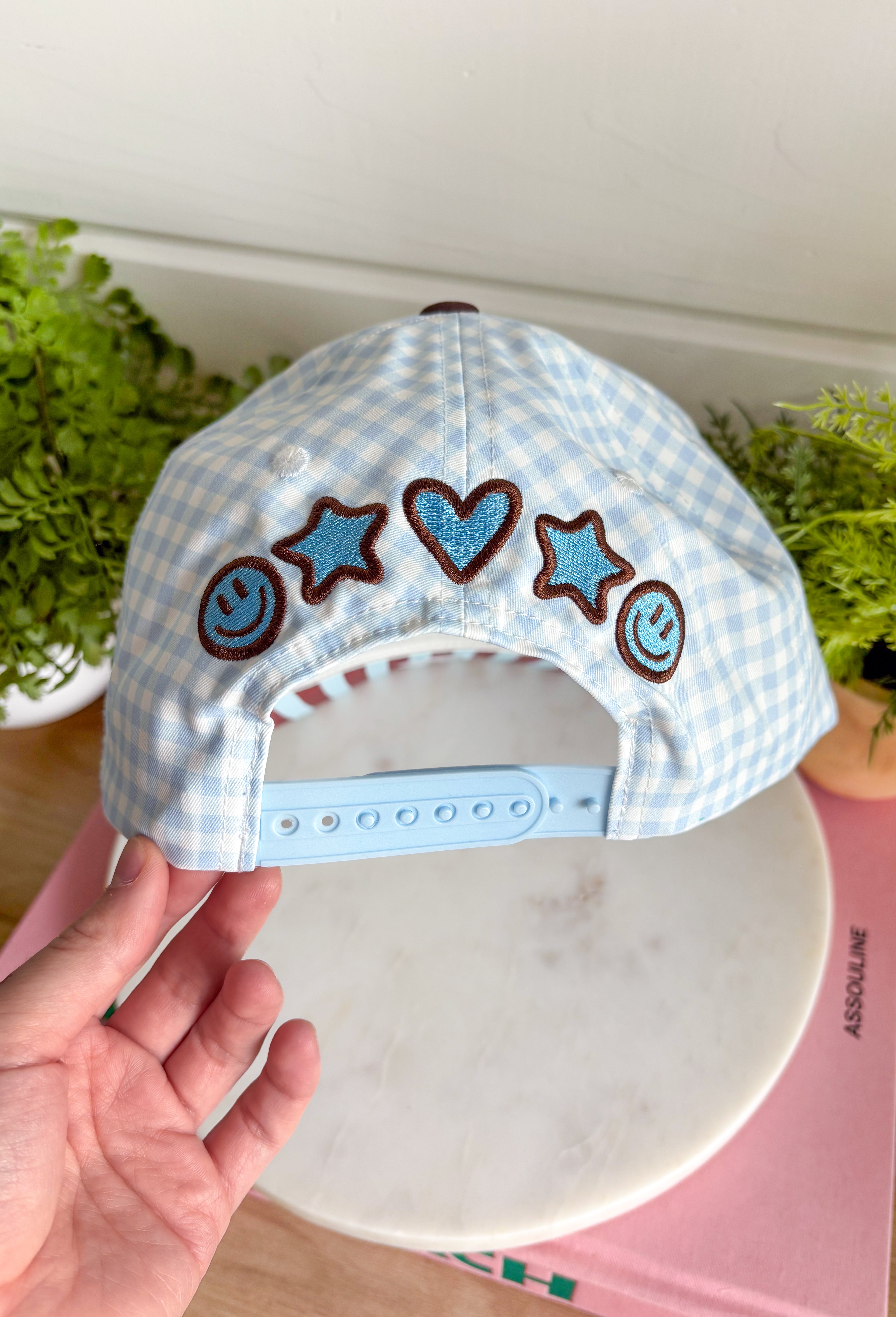 Bring a little fun to your everyday look with this charming trucker hat that's full of playful personality. Featuring a light blue and white gingham front paired with a light blue and brown striped bill, this unique hat is finished with white embroidered detailing along the bill for an extra touch of texture. The front is embroidered in chocolate brown with "Dilly Dally-er," while the side proudly reads "World's Best" with a ribbon. 