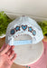 Bring a little fun to your everyday look with this charming trucker hat that's full of playful personality. Featuring a light blue and white gingham front paired with a light blue and brown striped bill, this unique hat is finished with white embroidered detailing along the bill for an extra touch of texture. The front is embroidered in chocolate brown with "Dilly Dally-er," while the side proudly reads "World's Best" with a ribbon. 